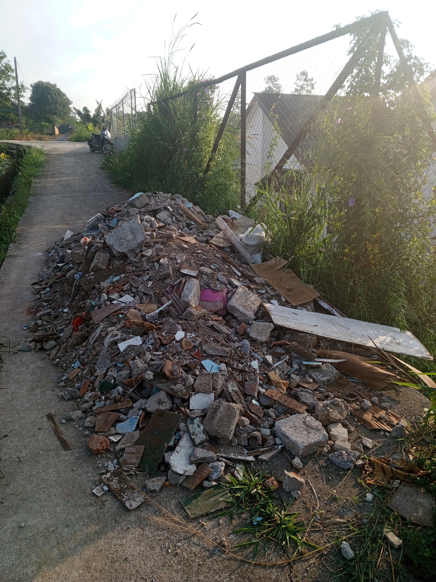 A bright outdoor scene features a large pile of construction or demolition debris on the side of a narrow concrete pathway. The debris, composed of broken concrete, tiles, bricks, wood fragments, and other rubble, is prominently positioned in the foreground to the right of the path. Fragments of blue and green tiles are visible among the predominantly gray and brown materials.
The concrete pathway extends into the midground, curving slightly to the left, with green grass and foliage bordering both sides. In the midground on the left, a dark-colored scooter is parked on the pathway. To the right of the debris pile, a tall metal fence with wire mesh is partially covered by dense, overgrown green vegetation, including tall grasses and bushes. Through the vegetation and fence, parts of a building with a pitched roof are visible in the background. The sun, casting a strong glare and lens flare from the right, illuminates the scene with a warm, bright glow, suggesting either early morning or late afternoon.
In the distant background on the left, large trees and hints of other structures can be seen under a clear sky. There are no people visible, and no active event is taking place, though the dumped debris implies prior activity. The setting appears to be a semi-rural or residential area, possibly a local track or service road. No discernible text is visible anywhere in the image.