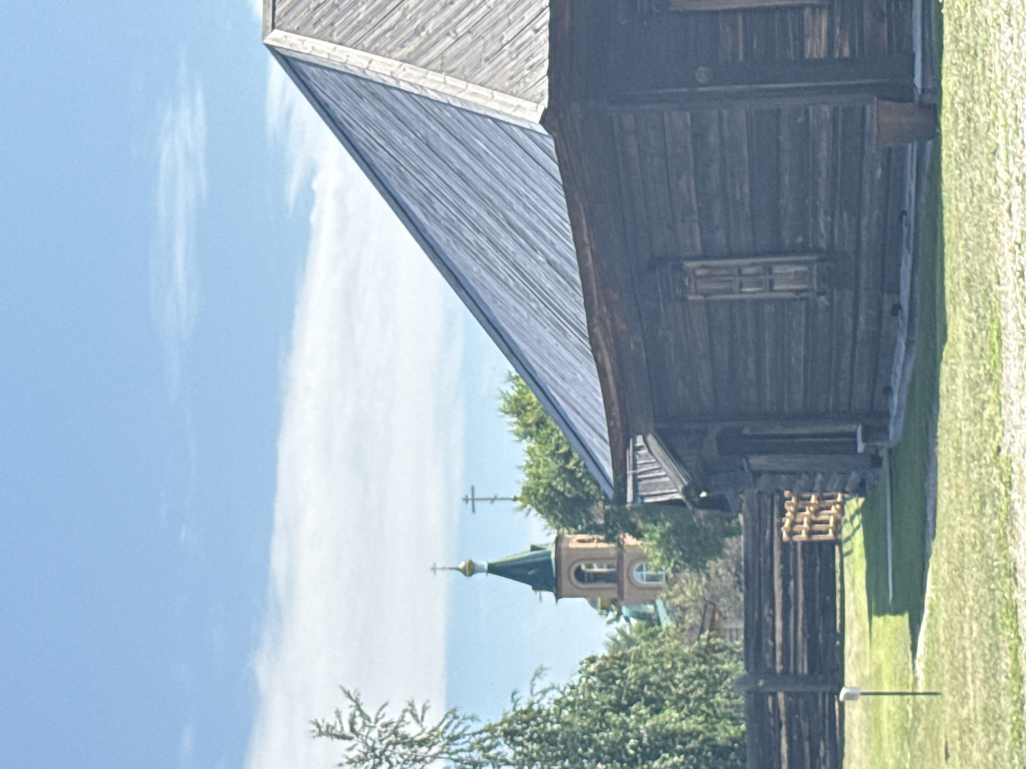 This is a vertical, outdoor, medium-distance shot of an old wooden structure with a metal roof in Shushenskoye, Russia. The wooden building is in the foreground and has a dark brown, weathered wood exterior with a small, shuttered window. To the left of the building is a weathered wooden fence. In the background, behind the fence and trees, is a Russian Orthodox church with a green steeple and a golden cross. The sky is blue with some white clouds. The scene is well-lit, suggesting it is daytime, likely sunny. The ground is covered with green grass.