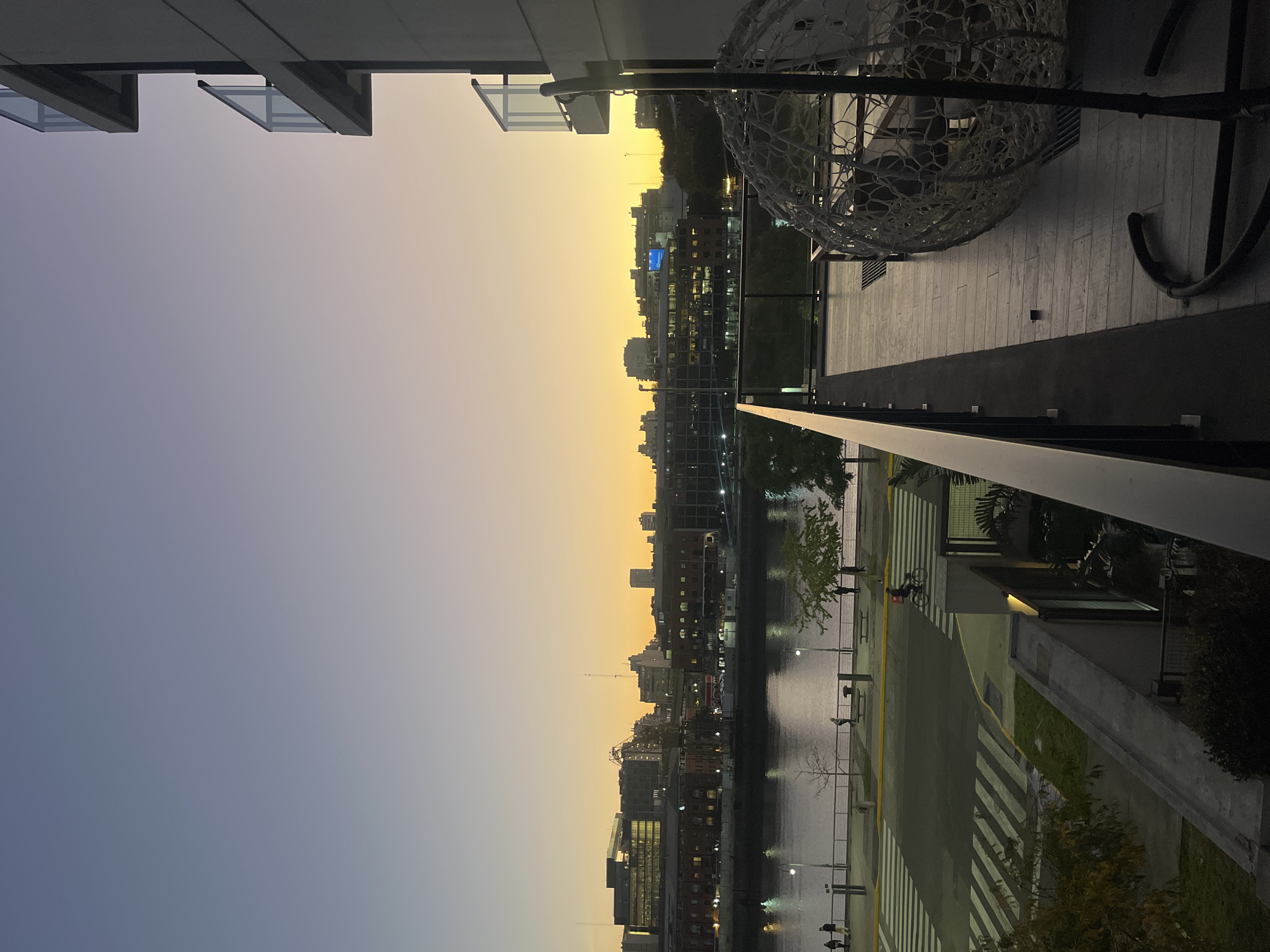 View from a balcony overlooking Puerto Madero in Buenos Aires, Argentina, during a sunset transitioning to twilight. The sky is a gradient of light blue to golden yellow. Buildings line the waterfront, some with visible lights on. The Río de la Plata is partially visible between the buildings. A black railing runs across the midground. On the balcony, a woven hanging chair is visible. The balcony has light-colored tile flooring.