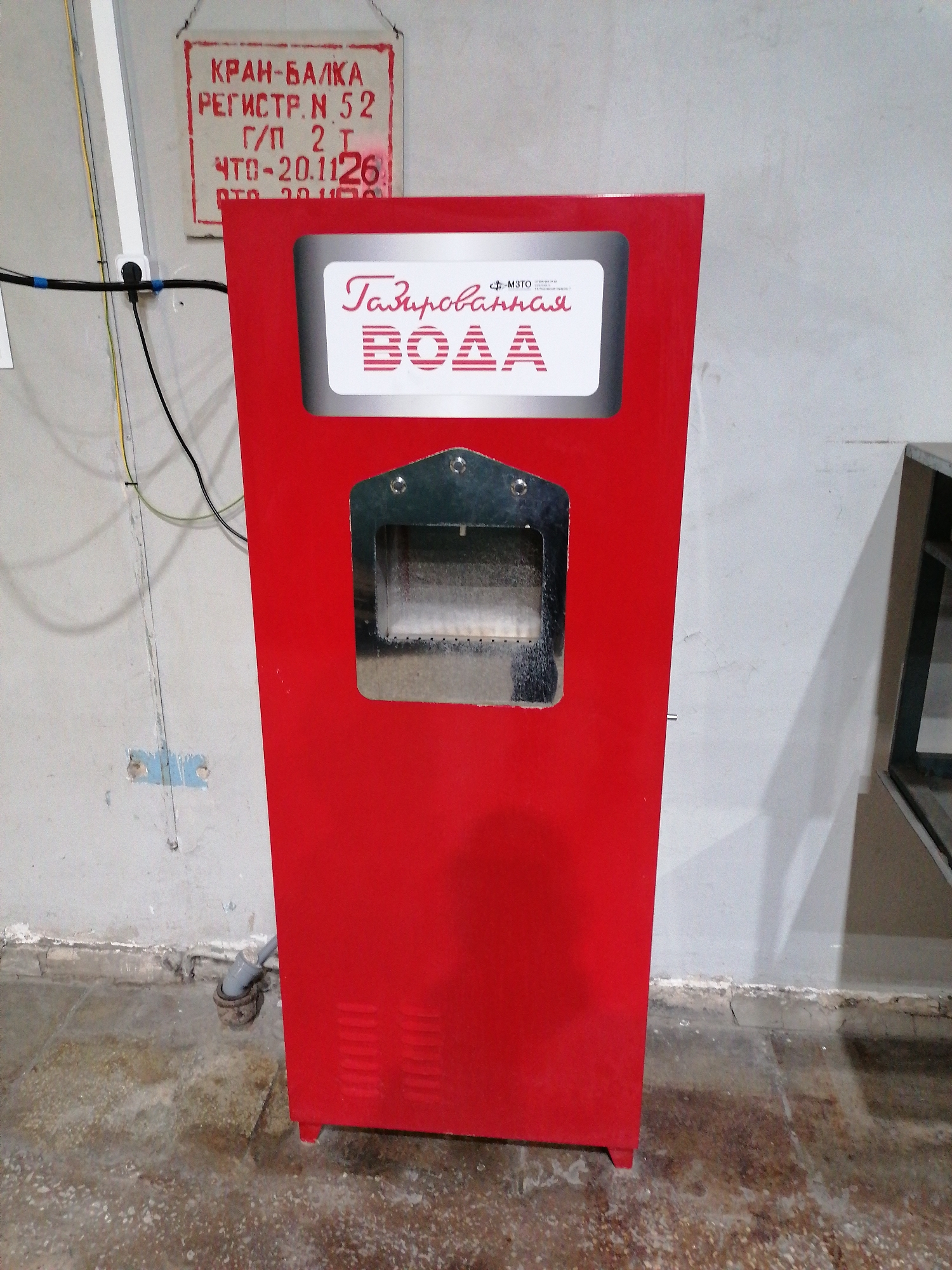 The image displays a bright red, freestanding vending machine for carbonated water, positioned against a light gray wall in what appears to be an indoor industrial or utility area. The vending machine features a prominent white panel at its top with the red Cyrillic text "Газированная ВОДА" (Carbonated WATER) and a small "МОТО" logo. Below this, there is a metallic, recessed dispensing area. Vents are visible on the lower right side of the machine, and a grey pipe exits its lower back, connecting to a drain on the floor.
The wall behind is a worn concrete surface, with electrical wires (black and yellow) running vertically and horizontally, secured by blue tape. A white rectangular sign with red text hangs on the wall, partially obscured by the machine. The visible text on the sign reads: "КРАН-БАЛКА" (Crane-Beam), "РЕГИСТР. № 52" (Register No. 52), "Г/П 2 Т" (Payload 2 T), and "ЧТО-20.1126" (likely a periodic technical inspection date).
The concrete floor beneath is dark, mottled, and appears wet or heavily stained. To the far right, a partial view of a dark, metallic structure is visible. The scene is static, unoccupied, and illuminated by artificial light.