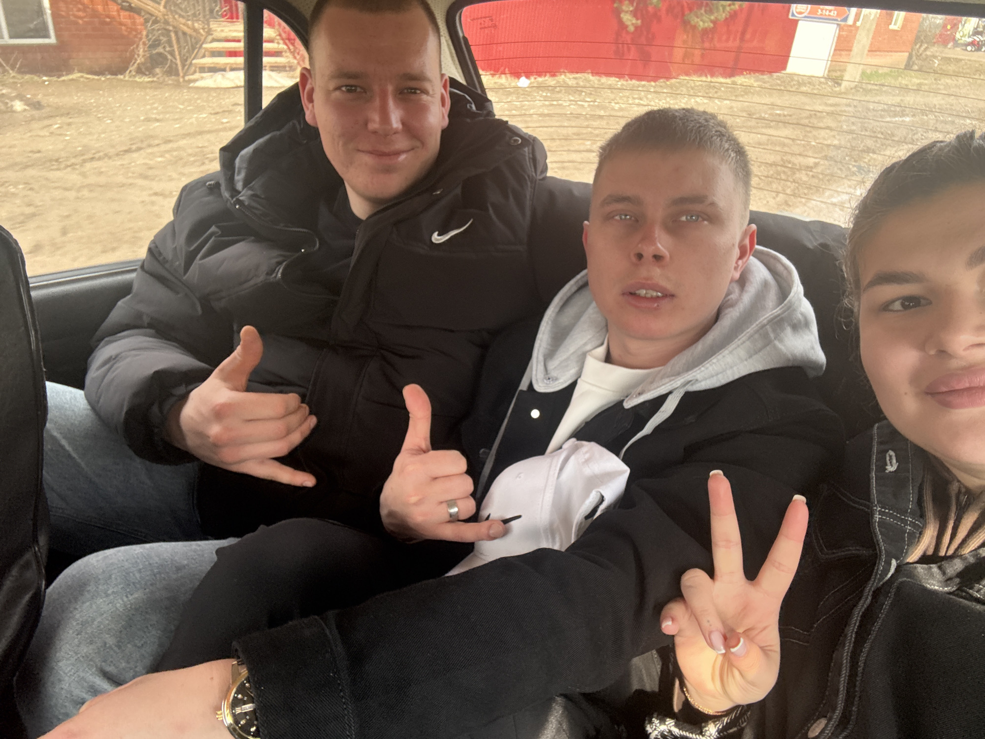 Three people are seated in a car. Two men sit in the back, while a woman is in the front passenger seat, partially visible. The man on the left wears a black puffer jacket and blue jeans and gives a "hang loose" hand gesture. The man next to him is wearing a grey hooded sweatshirt under a dark jacket and holds a white baseball cap. He is also giving the "hang loose" gesture, his middle finger extended instead of tucked. The woman in the front passenger seat raises a "peace" sign. The man to the left has a Nike logo on his puffy jacket. The woman has painted fingernails.
The background is blurry but visible through the car windows. A building is visible outside the window to the right, possibly a house. A building is also visible to the upper-right of the image, behind the back window. The sign 3-14-43 is visible on the building through the back window. Outside the window on the left is more of a rural setting.
