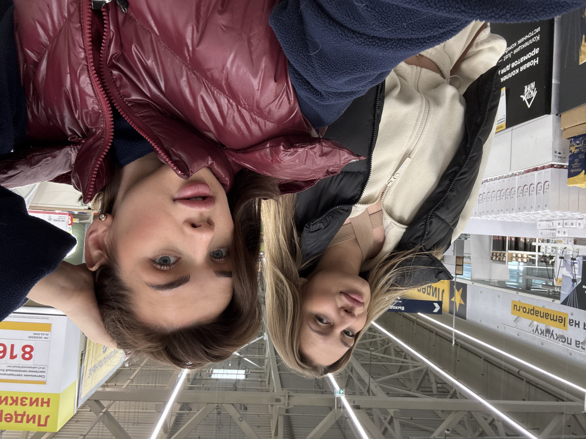 The image is an upside-down selfie taken by two women inside a retail store in Perm, Russia. The woman on the left has brown hair and blue-green eyes, wearing a burgundy puffer jacket over a dark blue fleece, with a small stud earring. The woman on the right has blonde hair, dressed in a cream-colored fleece under a black puffer jacket, with a beige top visible. Both women display neutral to slightly smiling expressions, suggesting a relaxed mood during their shopping trip.
The background shows the interior of a spacious store, possibly a supermarket or a home goods establishment, characterized by tall shelving, numerous products, and bright fluorescent strip lighting on the ceiling. Visible text, primarily in Russian, includes "Лидер" (Leader), "Низки" (Low), and an "Выход" (Exit) sign. A product label is visible, describing an "Светильник светодиодный настенно-потолочный Куб" (LED wall-ceiling light Cube) with specifications like "20 м, нейтральный белый свет" (20 m, neutral white light), a price or code "816", and an expiration or warranty date "25.03.2026". Other Russian phrases include "Новая коллекция ароматов для" (New collection of aromas for), "Коллекция Just" (Just Collection), "источник комфорт" (source of comfort), and a website "на lemanapro.ru". The English word "HOME" is also present on a product display. The image captures a casual moment of two individuals shopping.