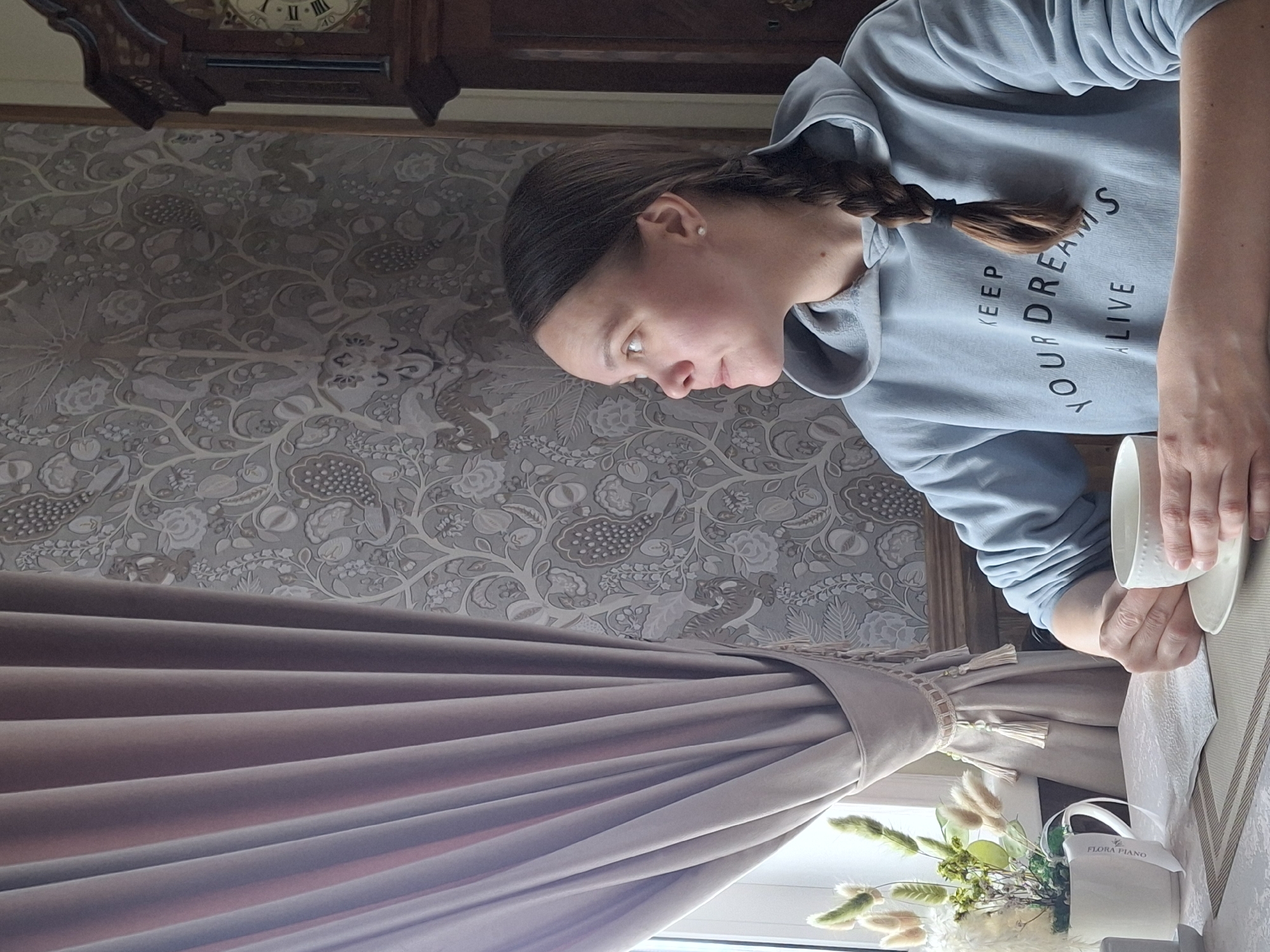 An indoor portrait shows a young woman from the chest up, focusing on her activity. She has light brown hair styled in a braid over her left shoulder and wears a light blue hooded sweatshirt with the black text "KEEP YOUR DREAMS ALIVE" printed on the chest. Her gaze is directed downwards as she holds a white ceramic mug and saucer with both hands, seemingly preparing a drink.
The background features a wall covered in elaborate patterned wallpaper with floral and possibly animal motifs in shades of grey and white. In the upper right corner, part of a dark wooden grandfather clock is visible. To the left, a heavy, light beige or taupe curtain is pulled back by a decorative tie-back with tassels and beads, revealing a window below. On a surface near the window, a white decorative pot, labeled "FLORA PIANO," holds a dried floral arrangement of fluffy white and green elements. The woman's hands and the items rest on a table or countertop, partially covered by a patterned textile. The scene is illuminated by natural light, suggesting it is daytime.