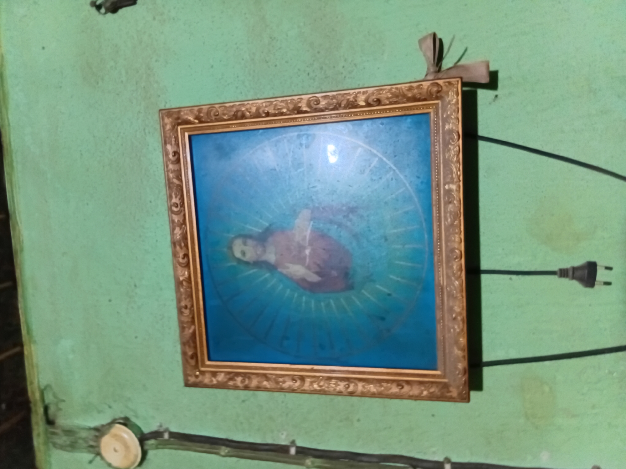 A framed portrait of Jesus is displayed on a green, textured wall. The portrait features a depiction of Jesus with long hair and a beard, wearing a reddish robe. He is positioned centrally within a blue background, which is adorned with radiating lines that create a halo effect. The frame of the portrait is ornate and golden. To the left of the portrait, an electrical conduit runs vertically down the wall, with a white circular fixture attached near the top. Below the portrait, a black electrical cord hangs, ending in a black plug. The wall appears to be an interior surface, possibly in a residence.