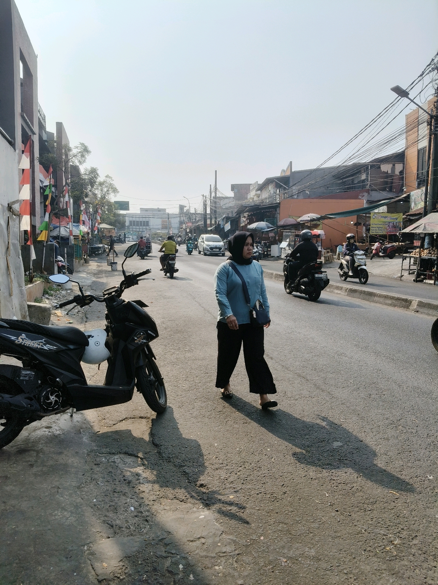 This is a medium shot, eye-level, natural light photograph of a street scene in South Tangerang, Indonesia. In the foreground, a black scooter is parked on the side of a paved road, casting a shadow. A woman wearing a blue long-sleeved shirt, black hijab, and black pants walks across the road from left to right. She has a blue shoulder bag. Behind her, traffic moves along the street, including motorbikes and a white car. Buildings line the right side of the road, with shops and stalls visible, some with awnings and merchandise displayed. Indonesian flags, red and white, are hung on poles along the left side of the street, near some buildings and trees. The sky is bright and hazy, suggesting it is daytime. The road appears to be in moderate condition with some cracks and uneven surfaces.