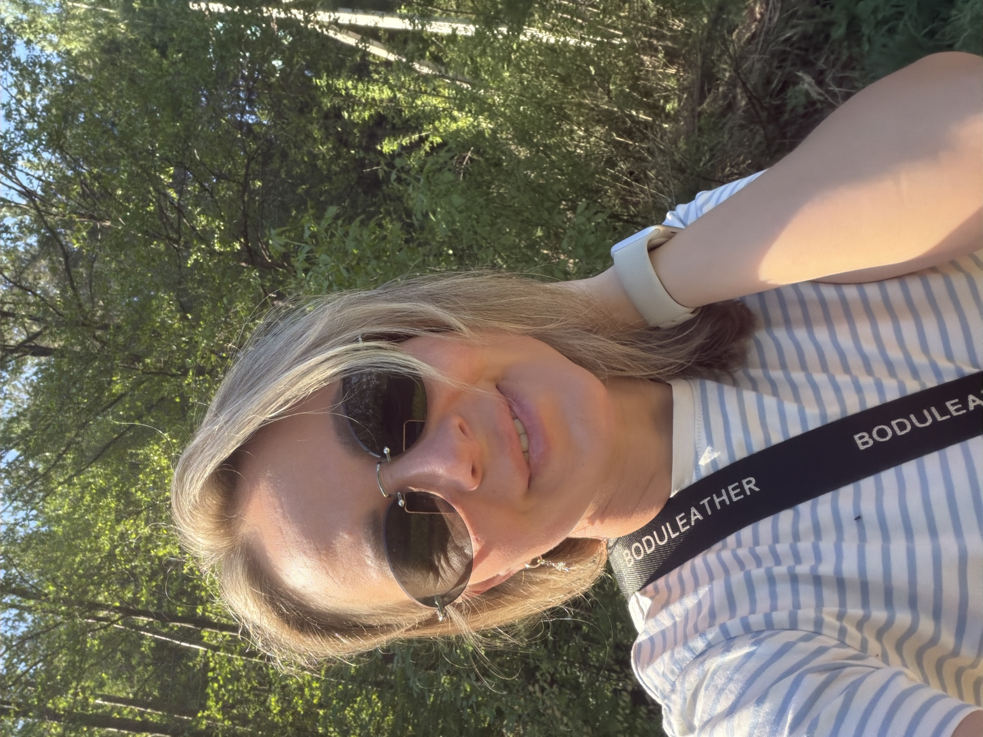 A woman with sunglasses is posing for a selfie. She is wearing a blue and white striped shirt with a strap that says "BODULEATHER" on it. She has a smartwatch on her wrist and is outdoors with trees in the background. The location is Chaikovsky, Russia.