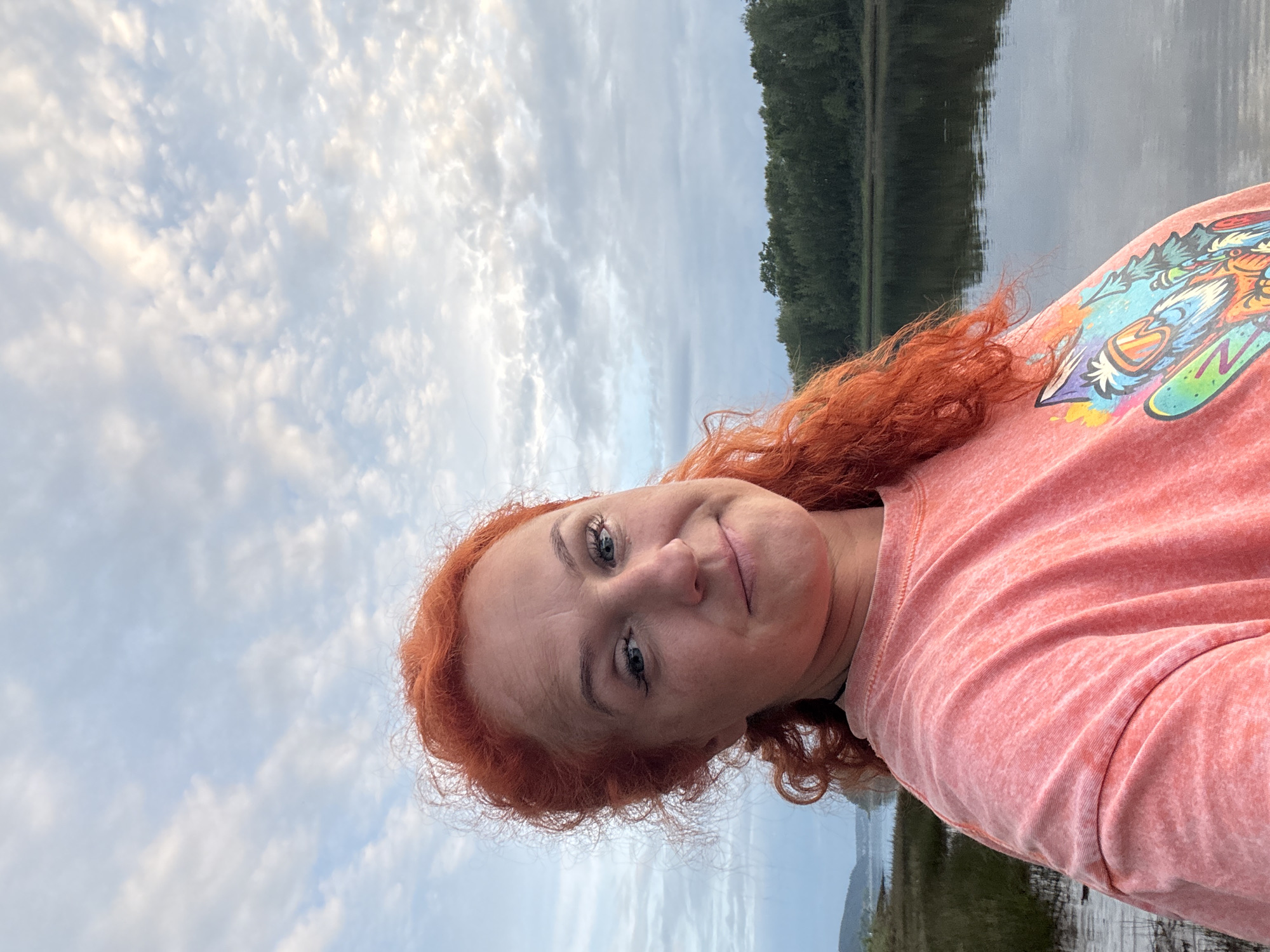 This is a rotated, head-and-shoulders selfie of a woman with vibrant red, curly hair. She is outdoors, with a body of water in the background, reflecting a treeline and the sky. The sky is filled with fluffy, white clouds, suggesting it is daytime, possibly late afternoon or early evening due to the soft lighting. The woman is wearing a coral-colored sweatshirt with a colorful graphic design on the left chest area, featuring cartoon-like characters with sharp features and bold outlines. Her expression is relaxed and she appears to be smiling gently. The overall impression is one of a casual, serene moment captured outdoors.