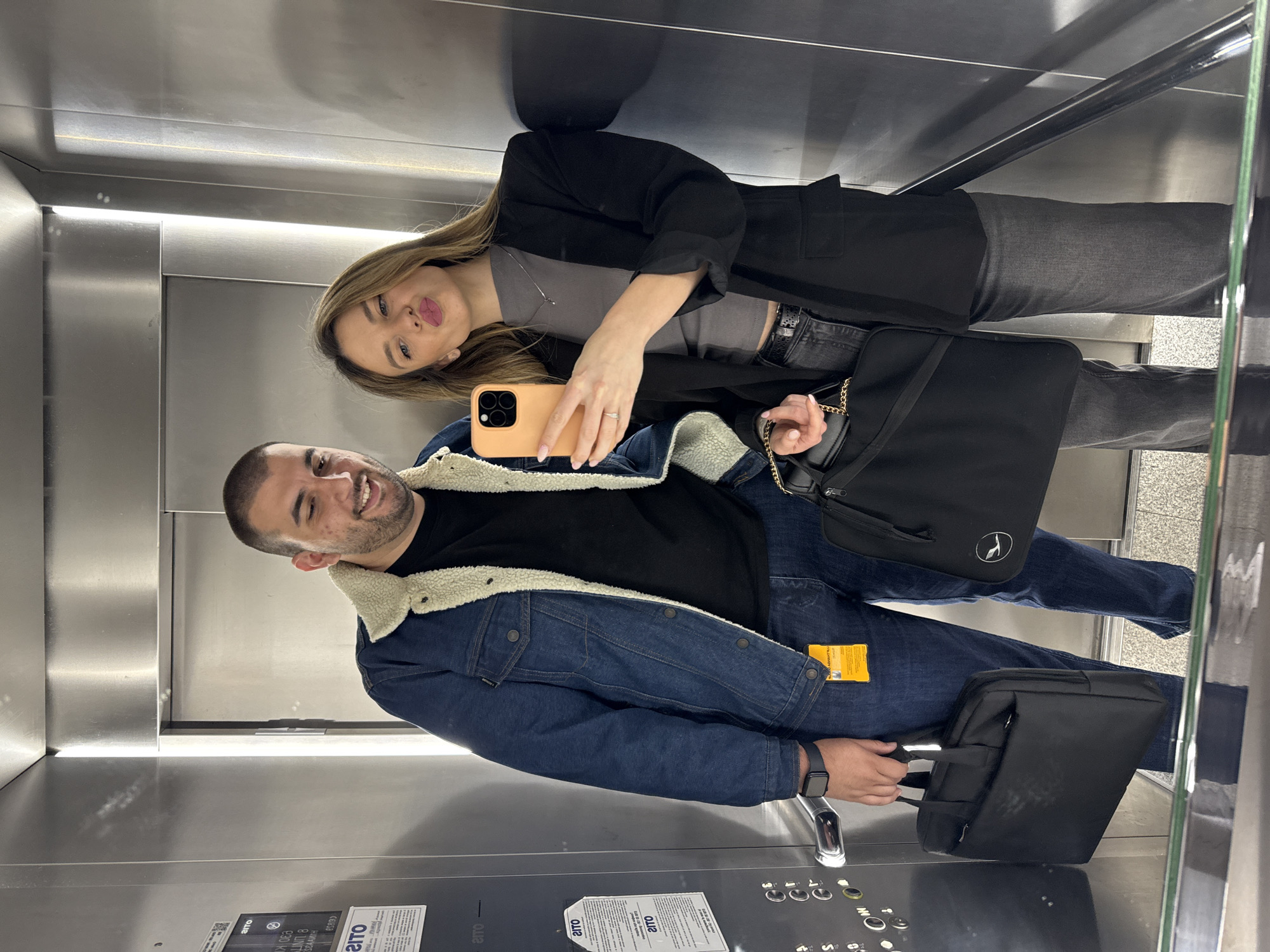 The image shows a man and a woman in an elevator, taking a selfie. The man is wearing a blue denim jacket with a cream-colored shearling collar, a black t-shirt, blue jeans, and a black smartwatch on his left wrist. He is carrying a black bag. The woman is wearing a grey top, a black blazer, grey jeans, and a ring on her left hand. She is holding a phone with an orange case to take the picture. She also has a black bag with a gold chain. The elevator has stainless steel walls and a control panel with buttons and informational posters. The location is in the city of Unknown, Unknown.
