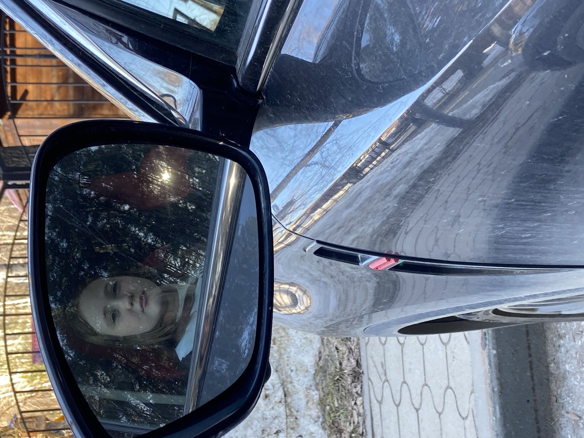 This vertical, outdoor photograph features a dark-colored car, likely black or dark grey, with a prominent side mirror in the foreground. The car appears somewhat dusty, with reflections of the bright sky and surrounding environment on its paintwork.
The primary subject is a young child, approximately 5-8 years old, reflected in the car's side mirror. The child has light hair and a neutral or slightly pensive expression, looking towards the viewer. They appear to be seated inside the car, likely in the rear or front passenger seat. A bright sun flare is visible in the upper right of the mirror, and reflections of trees and possibly a slatted fence are evident around the child's image. In the upper left corner of the mirror, a symbol resembling a heated mirror indicator (three wavy lines in a rectangle) is visible.
The car's side, including the door and front fender, shows distorted reflections of the environment. A small red and clear side indicator light is present on the front fender. The ground next to the car consists of light-colored, patterned pavement or a sidewalk, with some scattered dirt or sparse vegetation. Part of the car's front wheel and tire are visible at the bottom right. The bright sunlight indicates it is daytime with clear weather. The setting is an urban or suburban area in Chaikovsky, Russia.