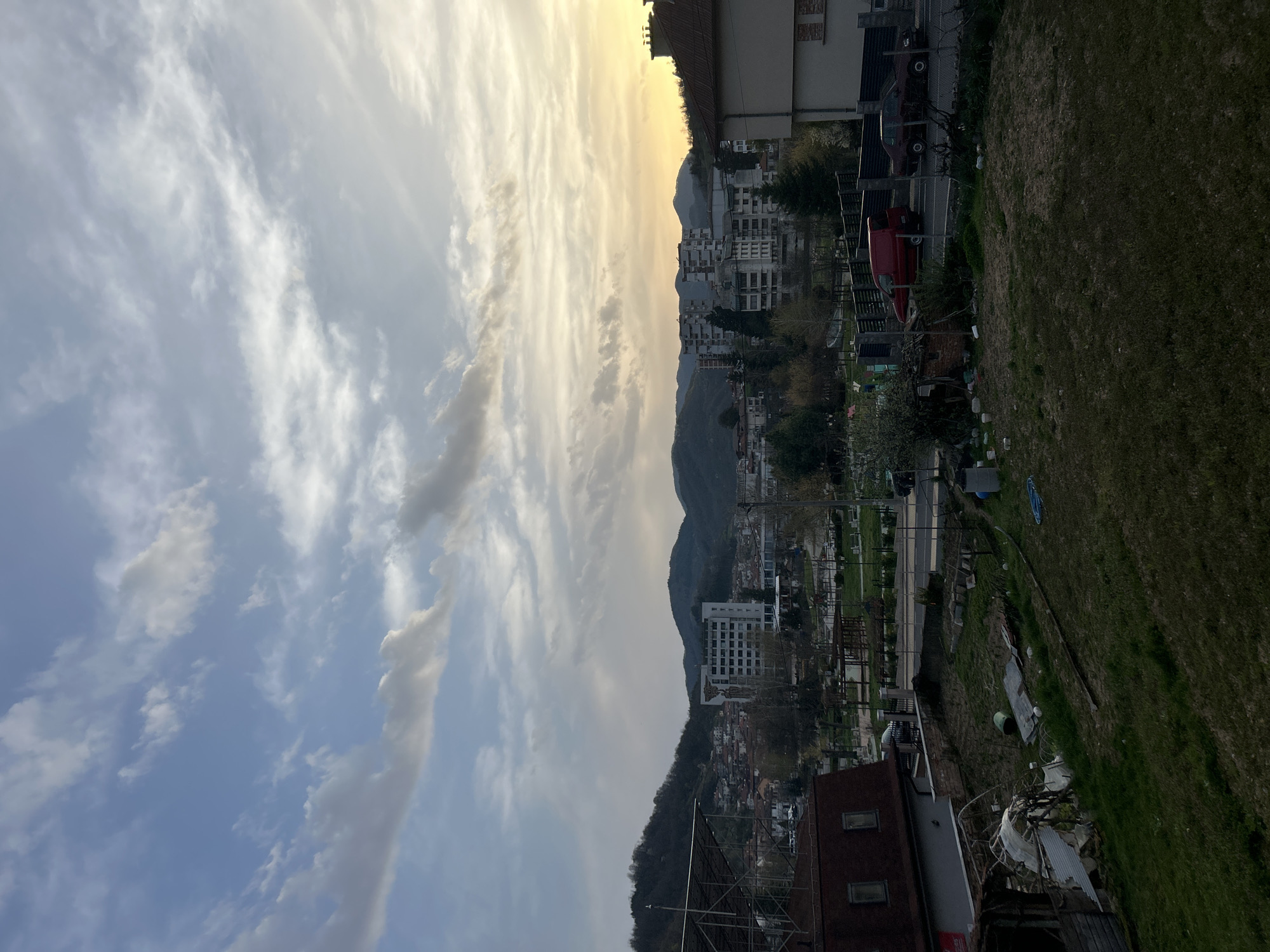 The image captures a cityscape of Zlatograd, Bulgaria, under a sky filled with scattered clouds and a hint of sunrise. The camera angle is slightly elevated, offering a panoramic view. Buildings of varying heights and architectural styles dot the landscape, nestled among hills and green spaces. A road or street is visible, suggesting urban development. The scene is quiet and still, devoid of any apparent human activity.