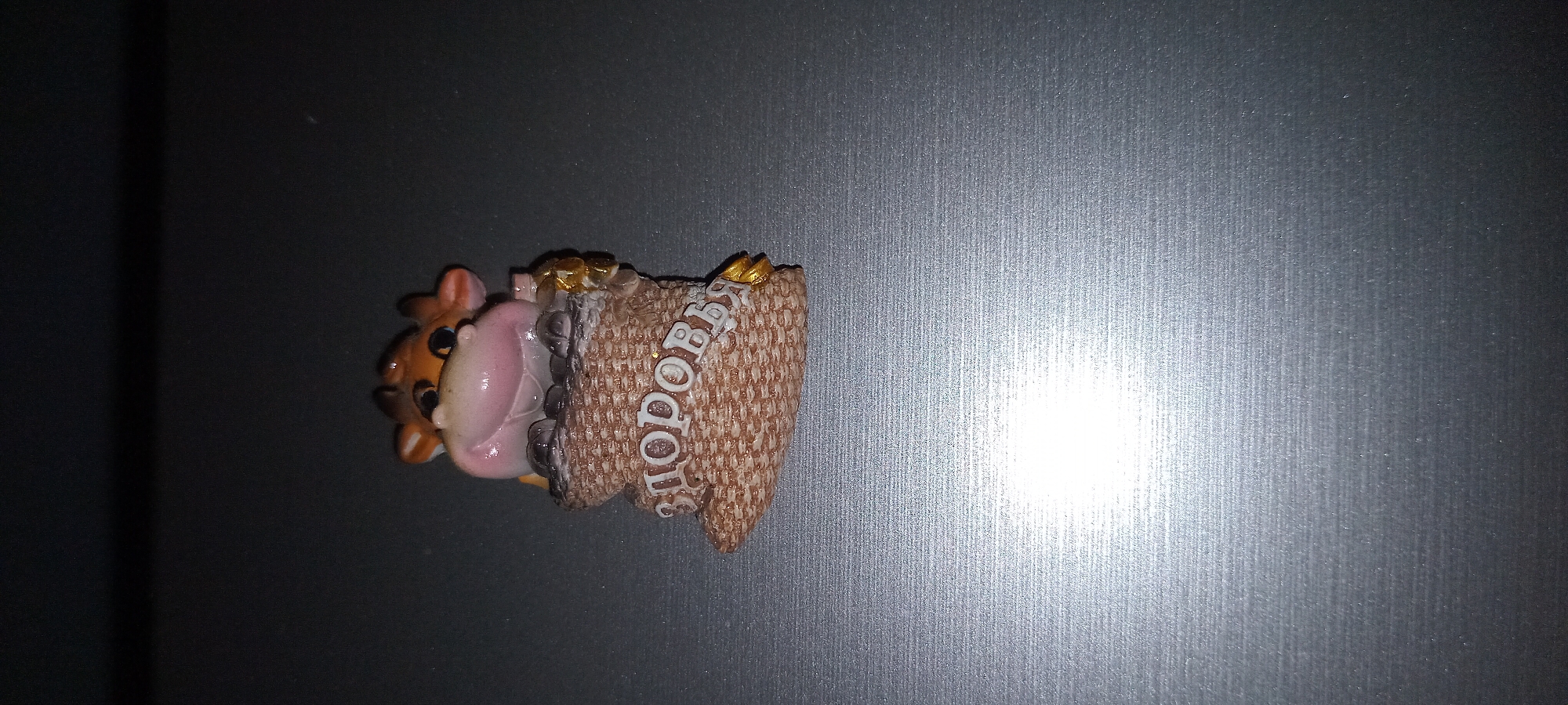 The image shows a small, whimsical figurine of a pig peeking out from a sack of money. The pig has a light pink body and a brown face with big, round eyes and a wide, open mouth with its tongue sticking out. It appears to be made of plastic or resin. The sack is textured to resemble burlap and is filled with what look like gold coins. The text "ЗДОРОВЬЯ" is written in white on the side of the sack. The figurine is placed on a dark, textured surface, possibly a refrigerator or a table, with a bright light source to the right creating a glare. The overall mood of the figurine is cheerful and suggests good fortune or wealth. There are no people visible in the image. The lighting suggests the photo was taken indoors, possibly in artificial light, given the distinct glare.