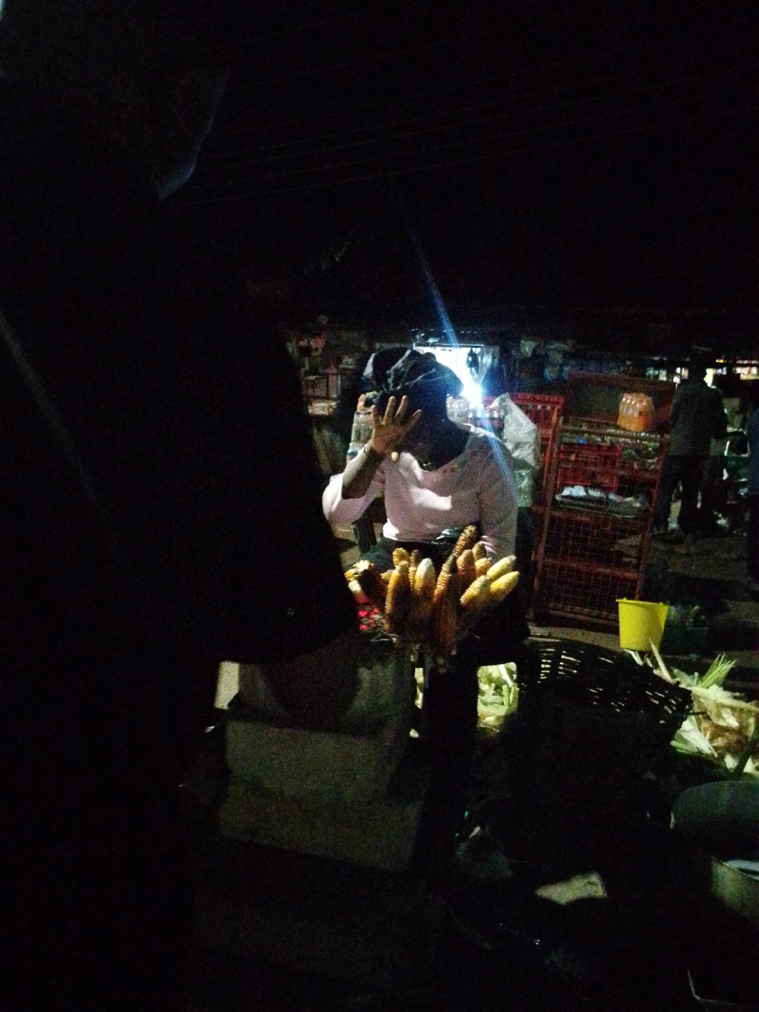 In a dimly lit outdoor market scene, a woman stands behind a display of corn on the cob. She is wearing a light-colored long-sleeved shirt and a headscarf, and her left hand is raised to her face, obscuring part of her forehead. In front of her, several ears of corn are arranged, some with husks still attached. The background of the scene is dark, with some faint outlines of structures, shelves, and other market stalls visible. A bright light source, possibly a lamp, illuminates the area around the woman and her corn, creating lens flare effects. A yellow bucket and other market items are scattered around. The overall impression is of a nighttime market in an urban setting, likely in Bukuru, Nigeria, given the context.