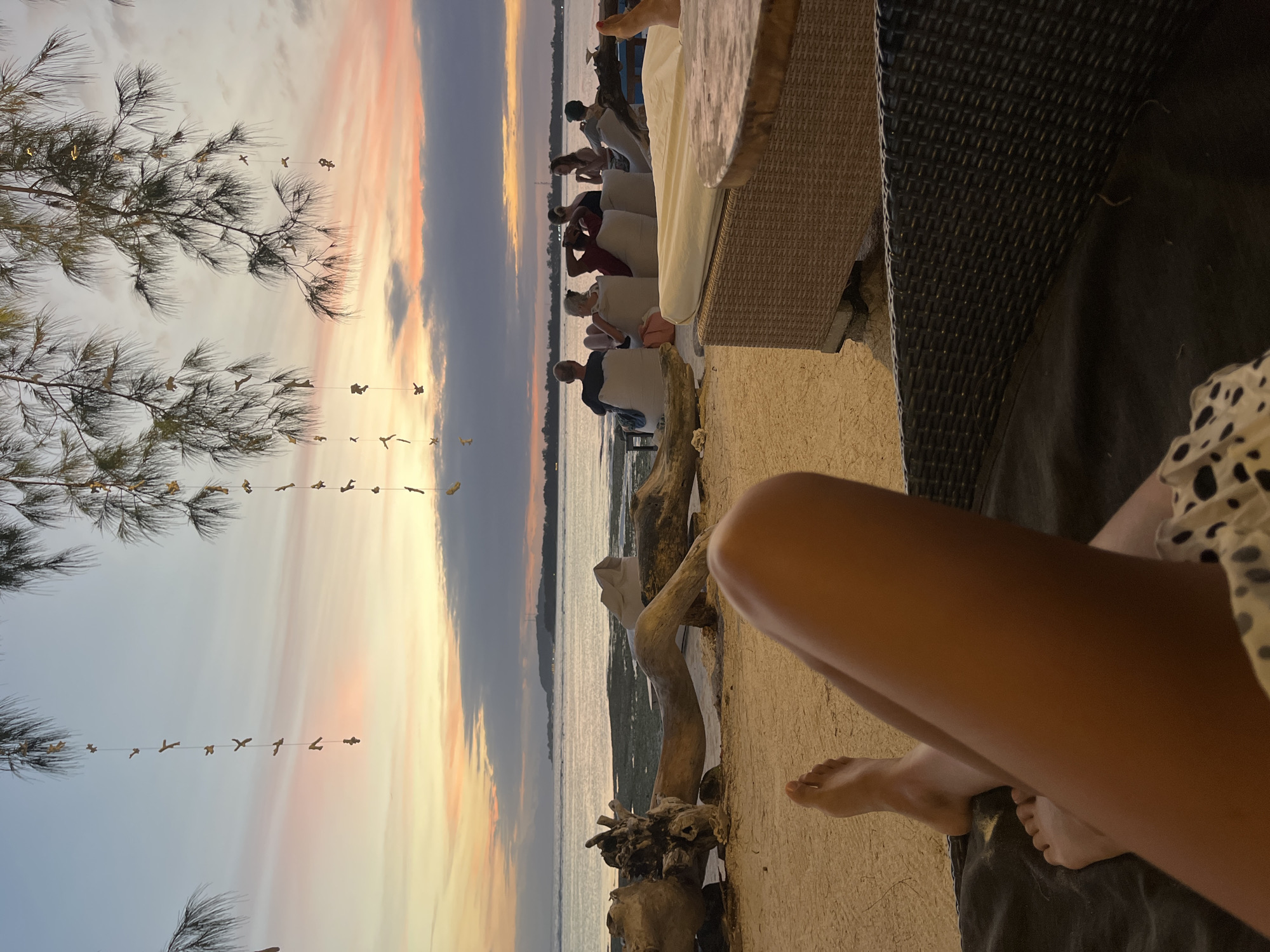 The image shows a sunset scene at a beach location, possibly a resort, in Unknown, Indonesia. A group of people are seated in the background, observing the sunset over the water. In the foreground, a person is seated with their legs visible; they are wearing a spotted print dress. A woven chair or lounge chair is also visible. Decorative driftwood sits on the beach sand. Strings of decorative objects hang from nearby trees. The sky shows a colorful sunset with hues of orange, pink, and gray.