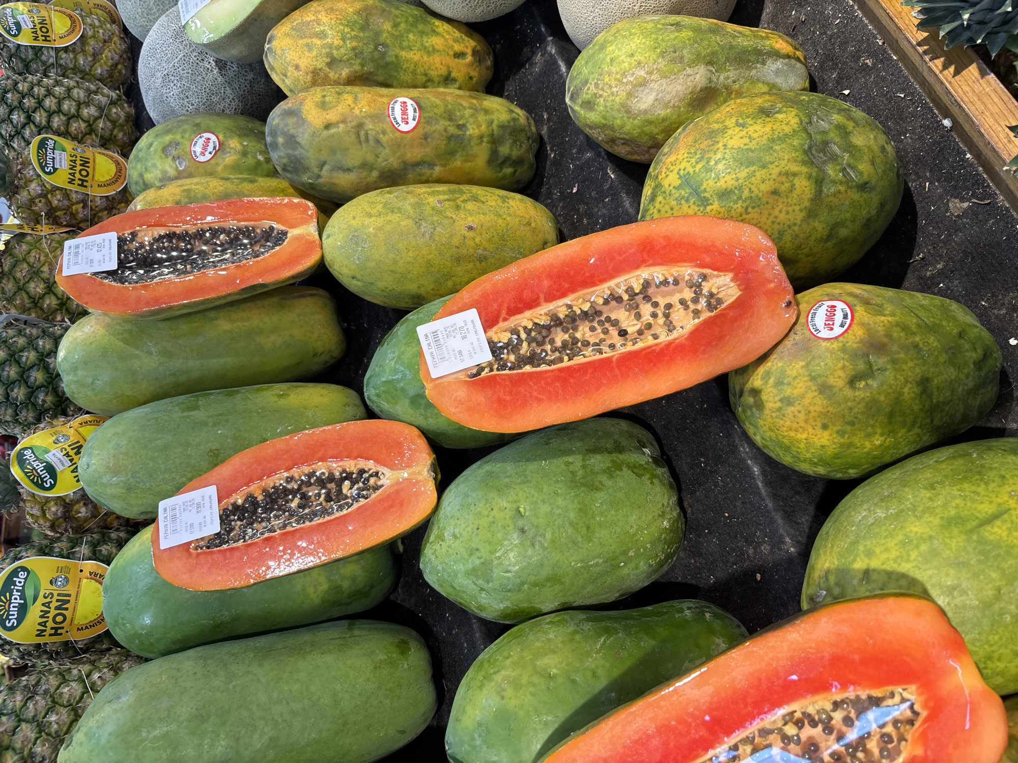 The image shows a close-up of a fruit stall in Kuta Selatan, Indonesia, displaying a variety of fresh produce. In the foreground, several ripe papayas are prominently featured, some whole and green, others halved to reveal their vibrant orange flesh and black seeds. One halved papaya has a price sticker labeled "PEPAYA CALIFORNIA." To the right of the papayas, a partially visible cantaloupe and other melons are present. In the background, pineapples with yellow "Sunpride" stickers are visible, indicating their brand and possibly origin. The stall appears to be set up outdoors, with a dark surface as the base for the fruits, possibly a market table or stand. The lighting suggests it is daytime, with shadows indicating the direction of the sun. The overall scene depicts a vibrant and abundant display of fresh fruits, typical of a local Indonesian market.