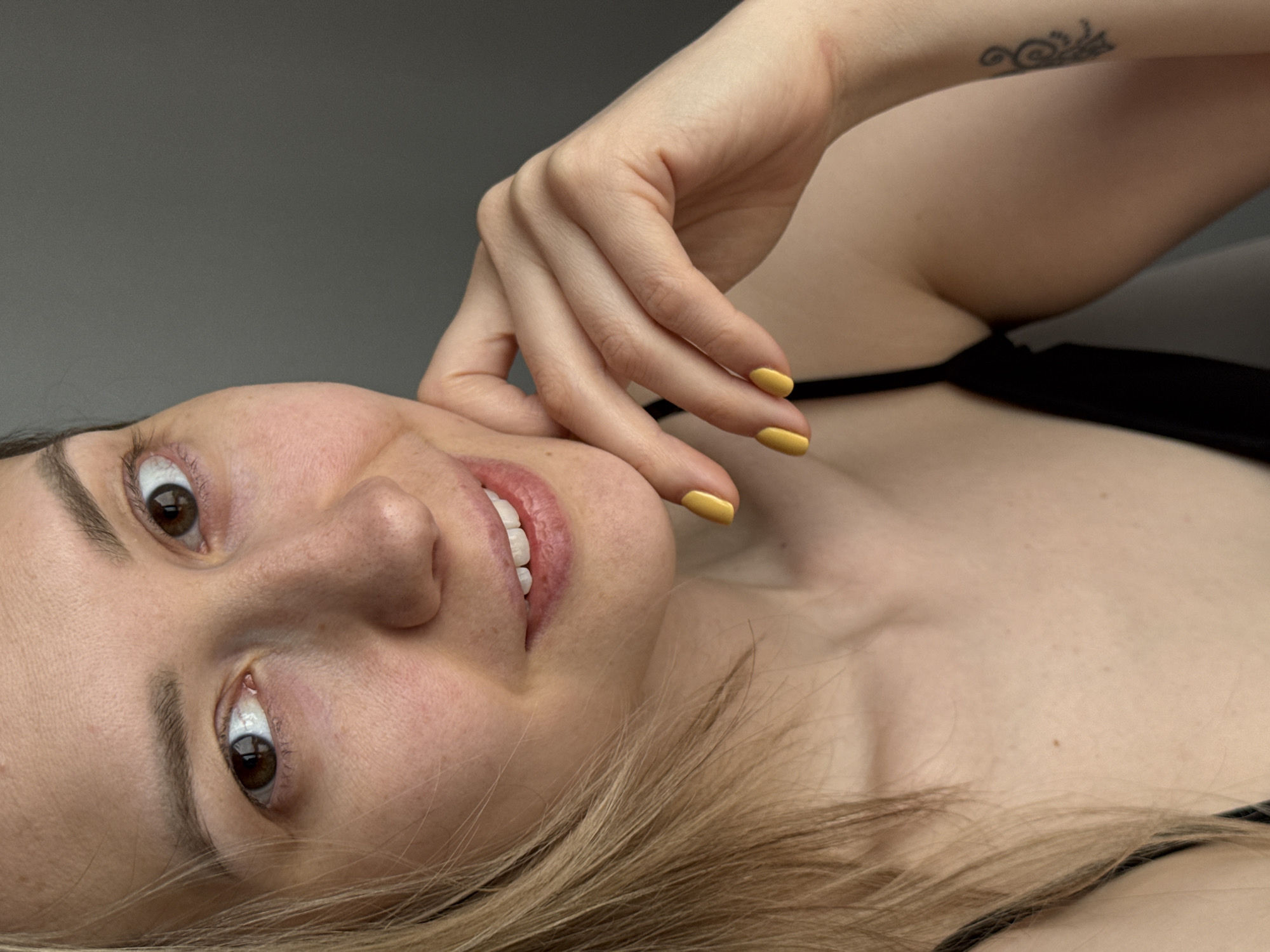 A close-up, tilted portrait shows a woman with fair skin and light brown hair partially obscuring her chest. She is lying down, looking at the camera with brown eyes and smiling slightly, revealing white teeth. Her right hand is raised, with her fingers near her cheek. Her fingernails are painted a bright yellow. She is wearing a black top with thin straps. A small tattoo is visible on her right forearm. There is no specific information to determine this image was taken in the city of Chaikovsky, Russia. The background is a neutral grey.