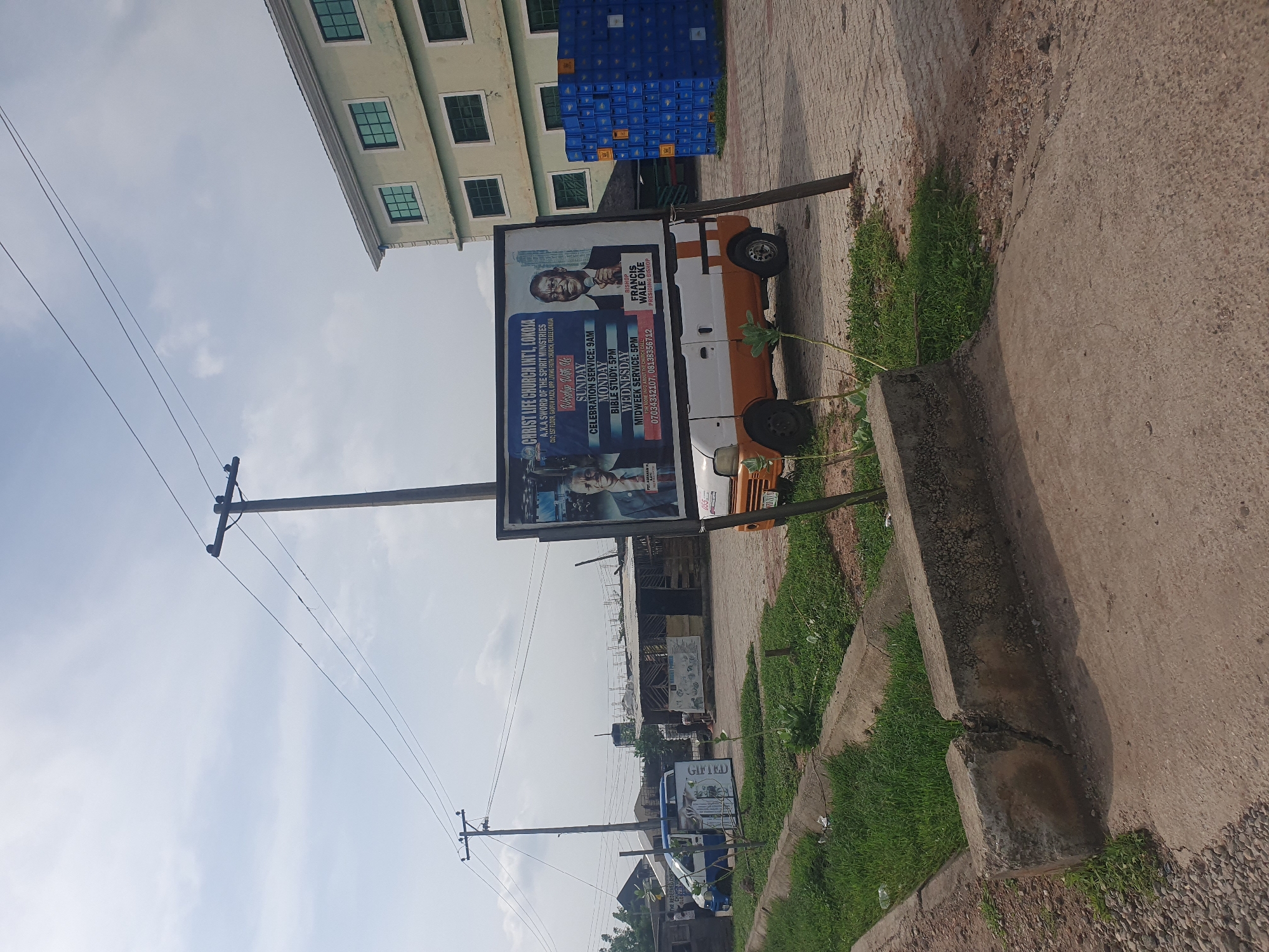 The image depicts an outdoor scene in Lokoja, Nigeria. A large billboard advertising "CHRIST LIFE CHURCH INT'L, LOKOJA" is prominently displayed. The billboard features images of two men, likely religious leaders, and text detailing service times: Sunday Celebration Service at 9 AM, Monday Bible Study at 5 PM, and Wednesday Midweek Service at 5 PM. Phone numbers are also listed. In front of the billboard, a white and orange van is parked. To the right of the billboard, a stack of blue crates is visible. Behind the van and billboard, there are low-rise structures, possibly shops or workshops, and a modern, multi-story building with several windows in the background. The environment includes a paved area, a grassy verge with a concrete curb, and overhead power lines supported by utility poles. The sky is partly cloudy. The image appears to have been taken during daylight hours.