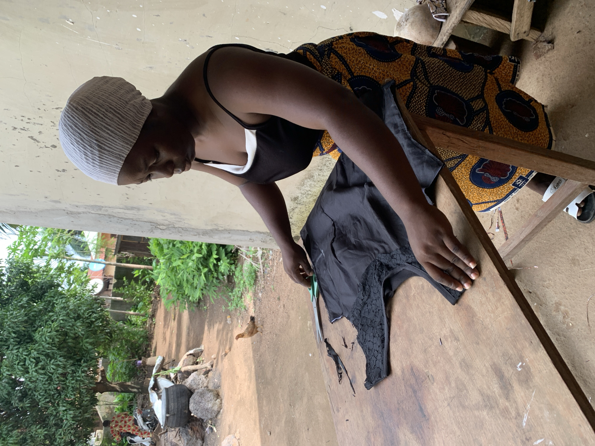 A woman is cutting black fabric on a wooden table in Akwanga, Nigeria. She is wearing a white headscarf, a black tank top, and a yellow and blue patterned skirt. Her hands are on the fabric, and a pair of green-handled scissors is visible. In the background, there is a wall, greenery, and a chicken.