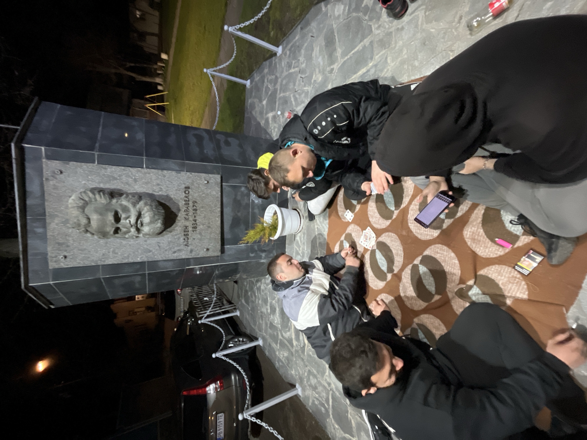 At night, a group of people are gathered on a brown patterned blanket next to the memorial of Lyuben Karavelov in Karavelovo, Bulgaria. They are playing cards. One person is looking at a smartphone. A flower pot sits near the memorial. A parked car is visible in the background. A clear bottle is on the stone pavers.