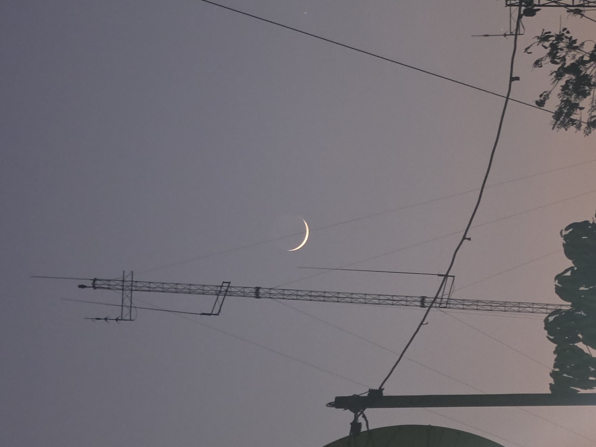 A nighttime scene in Pintomakmur, Indonesia shows a crescent moon in the sky above an urban environment. The sky is a dusky gray. An antenna tower and overhead power lines are silhouetted in the foreground. Plant life is present on the right side of the frame.