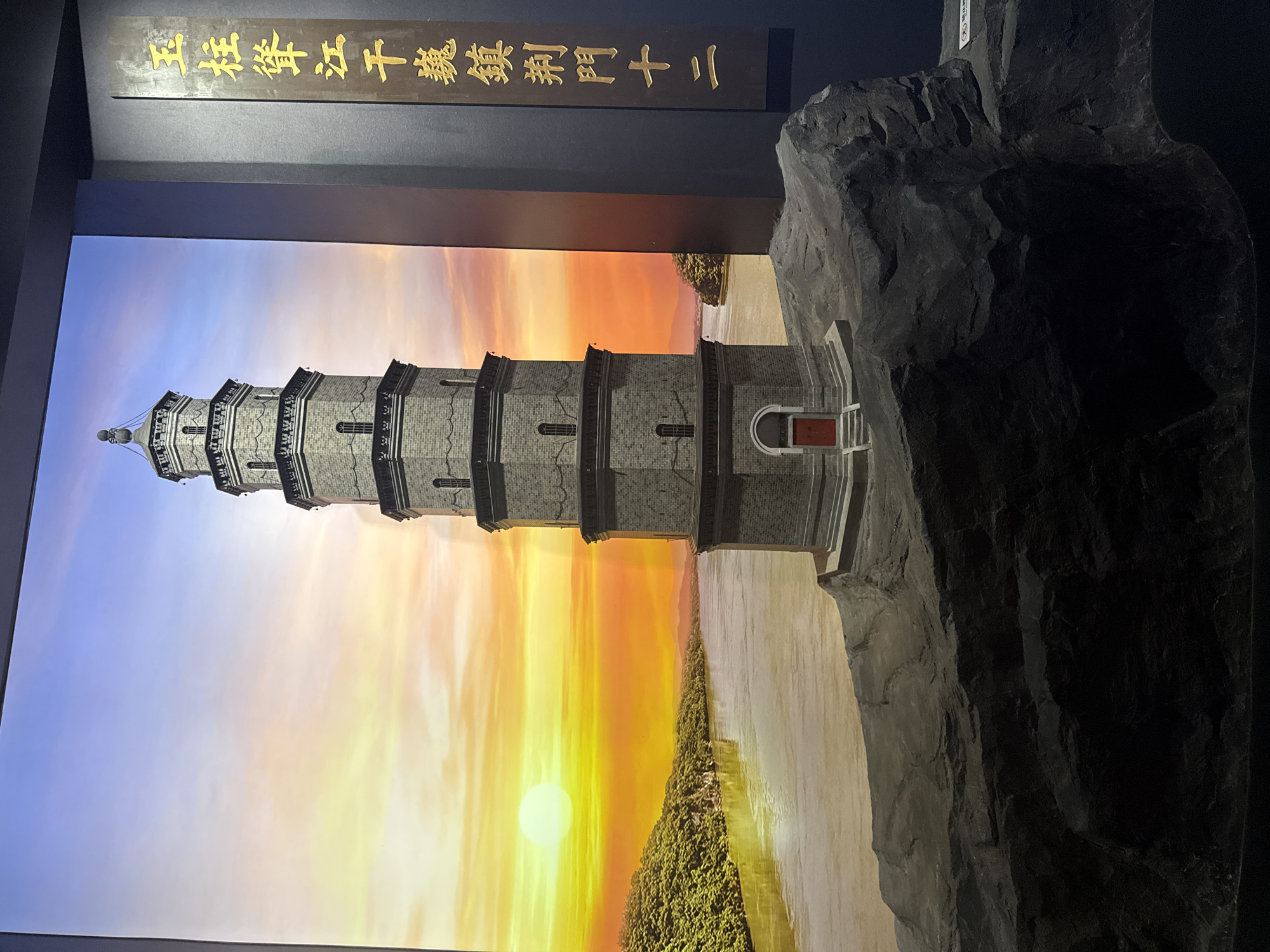 A model of a multi-tiered pagoda stands on a rocky outcropping. The pagoda is constructed of grey bricks and has a red door at its base. Behind the pagoda, a painted backdrop depicts a vibrant sunset over a river and a tree-covered shoreline. To the right of the pagoda, a dark vertical plaque with gold Chinese characters is visible. The scene appears to be an exhibit, possibly in a museum, showcasing a historical landmark. The overall impression is one of a diorama designed to recreate a specific landscape and architectural feature. The text on the plaque may indicate the pagoda's location or significance, possibly within Yuzhong District, China, given the context of the prompt. However, the image itself does not explicitly confirm this location.