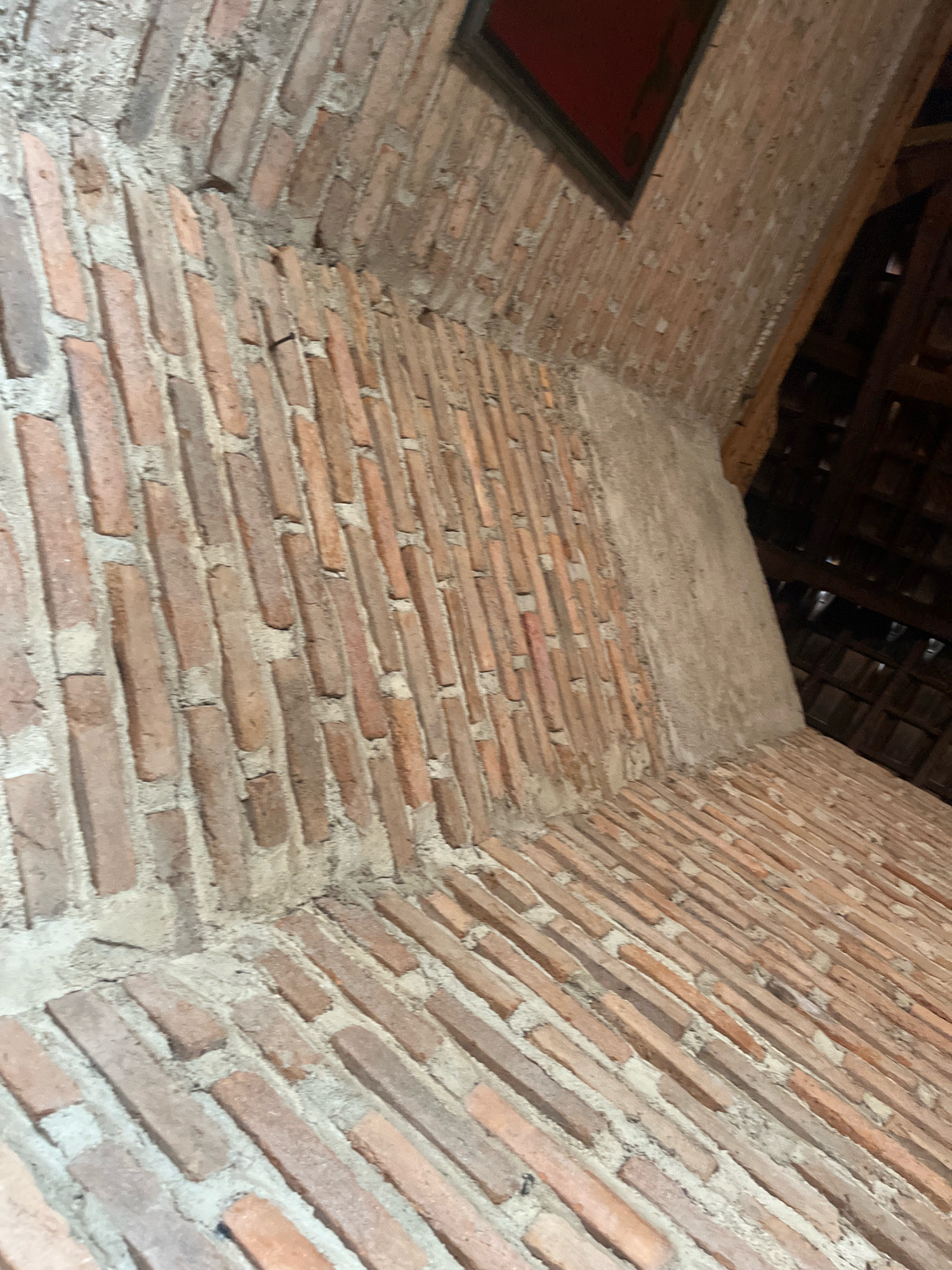 The image shows an interior view of a brick structure with a distinct arched ceiling and a floor laid with bricks. The bricks are a reddish-brown color and appear to be old and weathered, with visible mortar joints. A section of rough, gray plaster is visible on the curved wall. In the upper part of the image, a dark-framed opening with a red interior is visible. The background suggests a roof structure with wooden beams and tiling. The perspective is from a low angle, looking upwards and across the room. There are no people or animals in the image. This scene is located in the city of Sidomulyo, Indonesia.
