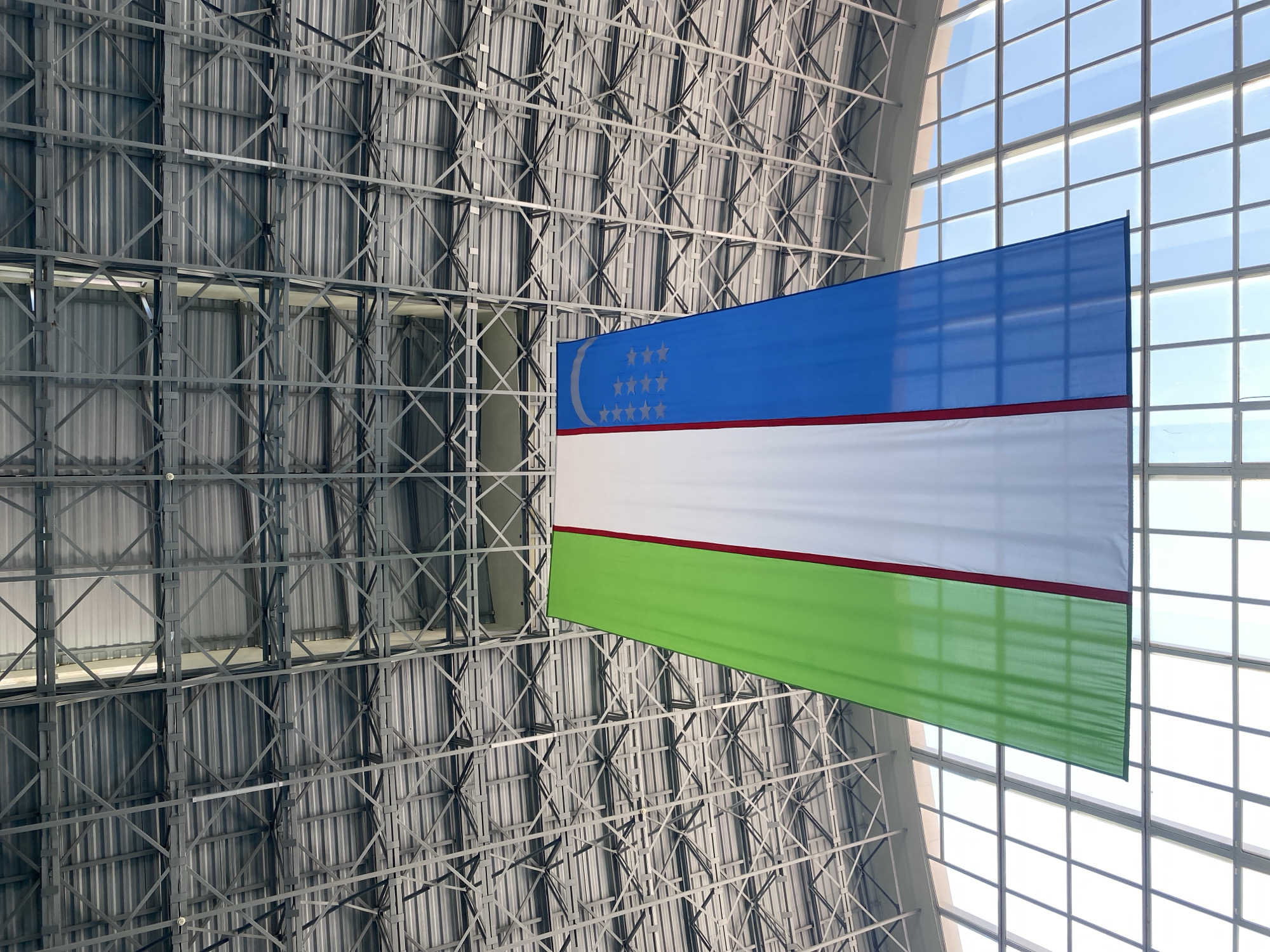 The image shows a large Uzbek flag hanging from a high ceiling structure in what appears to be a public building, possibly an airport or a convention center. The flag is vertically divided into three equal bands of light blue, white, and green, separated by thin red stripes. The blue band on the right features a crescent moon and twelve stars, symbols of Uzbekistan's independence and spiritual heritage. The ceiling is made of a complex metal framework supporting corrugated metal sheeting, and large windows with a grid pattern are visible in the background. The lighting suggests an indoor setting with natural light entering through the windows. The scene is devoid of people and any specific contextual clues linking it to Gurlan, Uzbekistan.