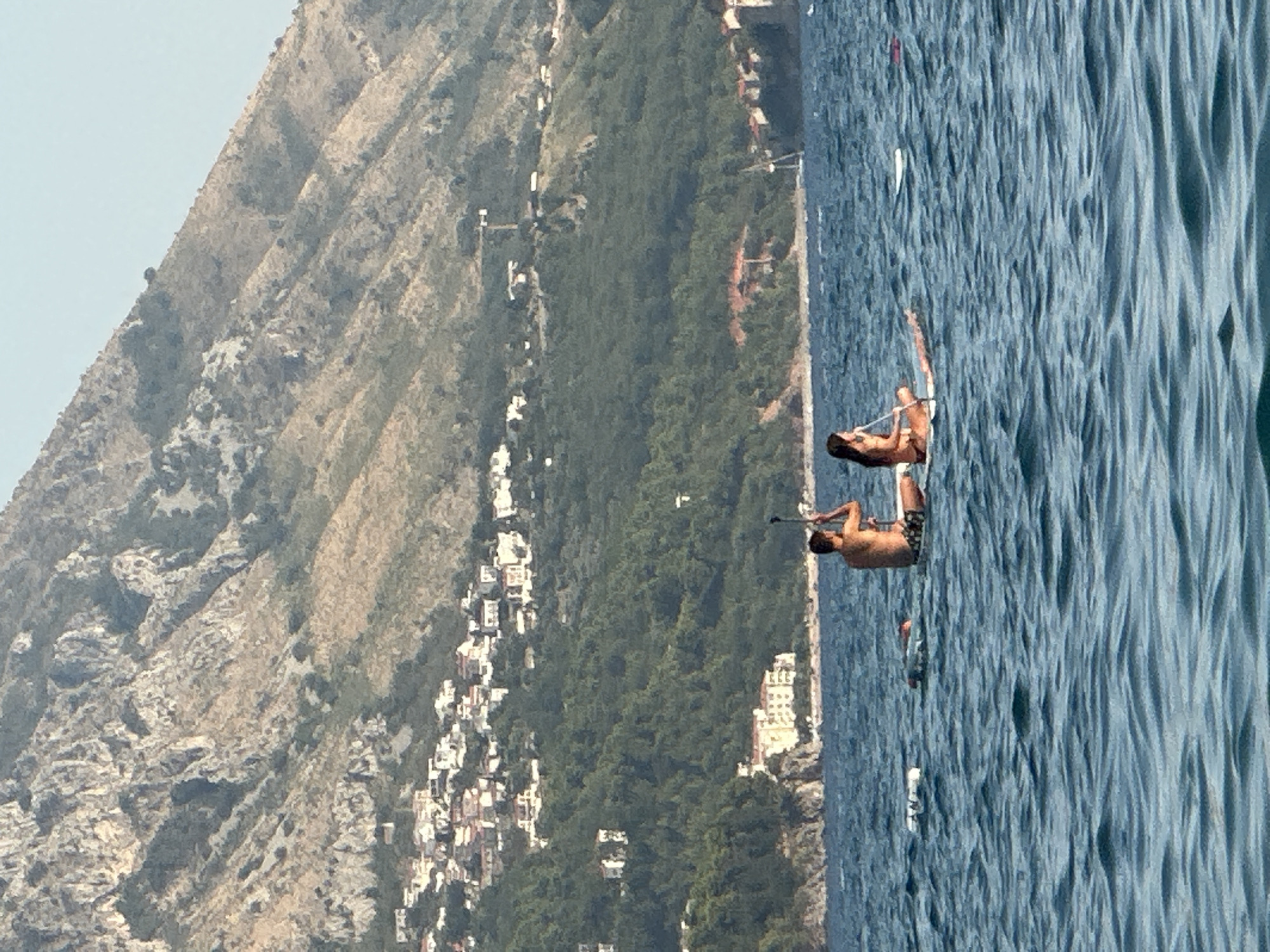 Two people are paddleboarding on a body of water with a mountain and a town in the background. The person on the left is a male wearing patterned shorts. The person on the right is a female wearing a bikini. They are both sitting on their paddleboards and holding paddles. The water is blue and choppy. The mountain is steep and rocky, with vegetation and buildings on its slopes. The town is clustered on the hillside. The image appears to be taken in Boreti, Montenegro, a coastal town known for its scenic beauty.