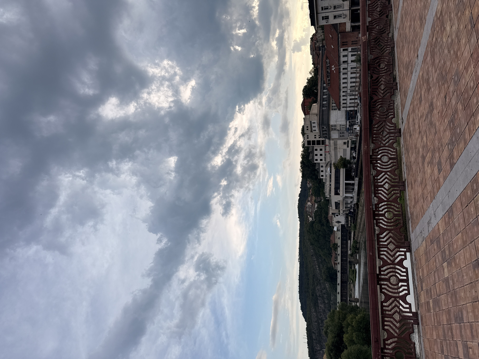 The image depicts a view from a bridge in Lovech, Bulgaria, overlooking the Osam River and the city's historic architecture. The foreground features a pedestrian walkway paved with reddish-brown bricks and a decorative metal railing. Beyond the railing, the Osam River flows, with reflections of the sky and surrounding buildings visible on its surface. Across the river, the city of Lovech unfolds, characterized by a mix of modern and older buildings. Dominating the skyline are the hills of Lovech, covered with lush green trees and dotted with houses. In the distance, a covered bridge, a prominent landmark of Lovech, spans the river. The sky above is filled with dramatic, grey clouds, suggesting an overcast or potentially stormy weather condition. The overall scene captures a scenic urban landscape with a strong sense of place, highlighting the natural beauty and historical architecture of Lovech.