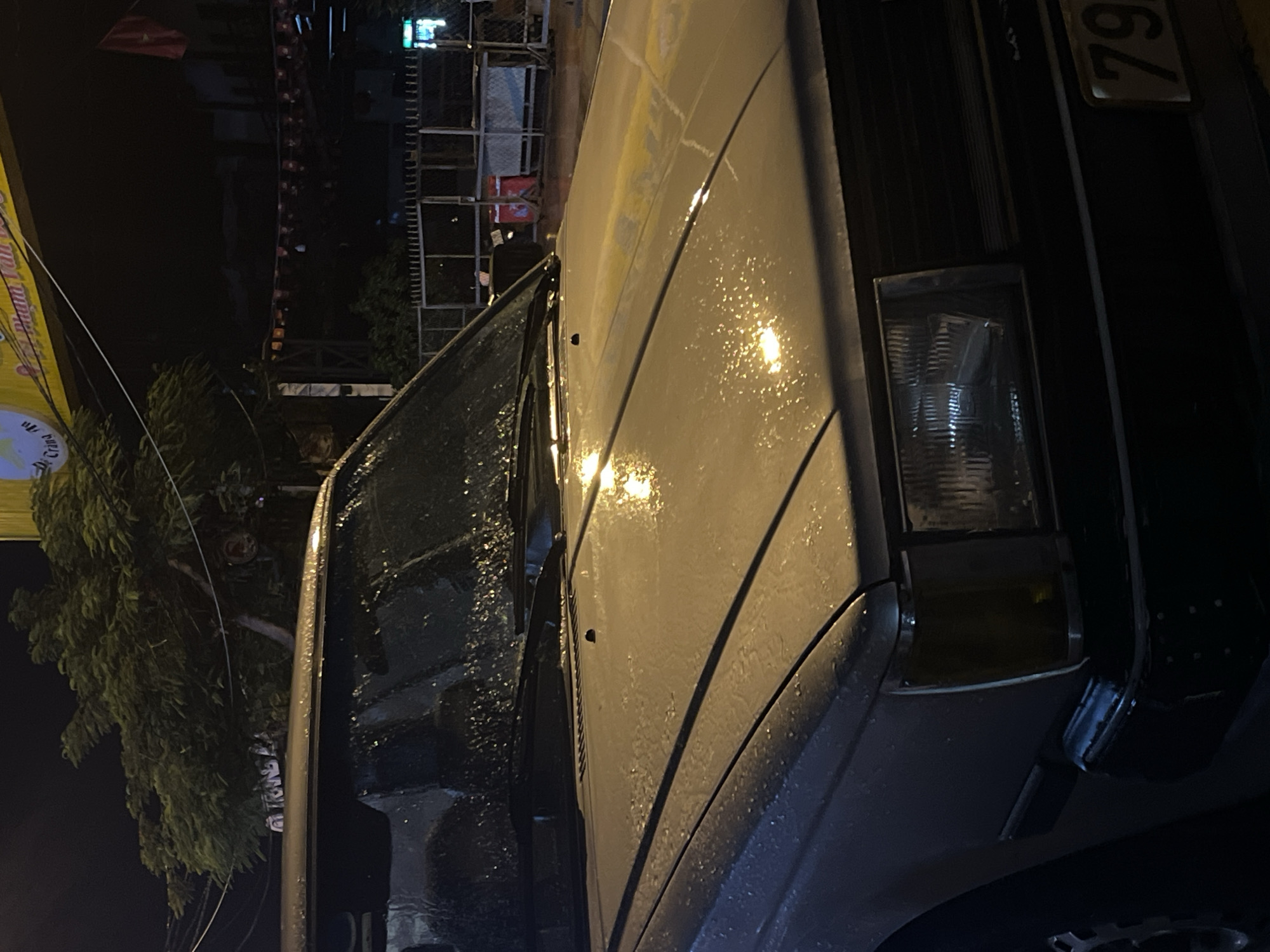 A vehicle with the license plate "79" is parked in Bac Nha Trang Ward, Vietnam, presumably at night. Rain is falling. A structure with scaffolding is visible behind the vehicle. A building with a yellow banner is in the background. A tree is on the left.