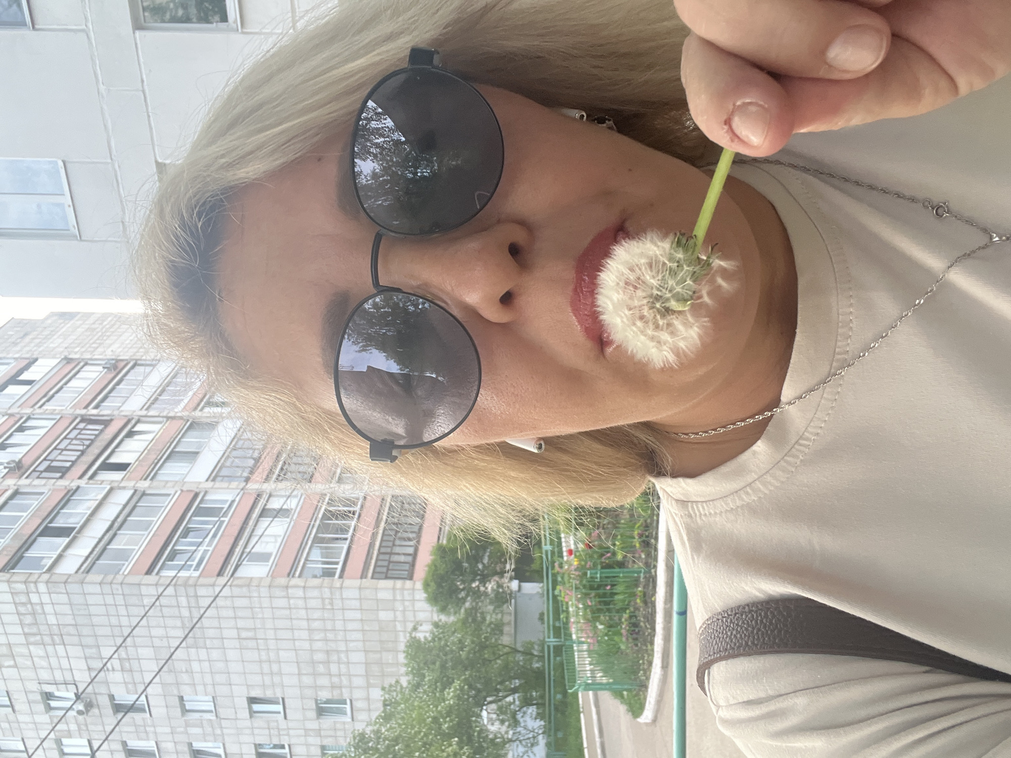 A woman with blonde hair and sunglasses is holding a dandelion in front of her face. She is wearing a white t-shirt and a silver necklace. There is a building in the background. The location is likely in Chaikovsky, Russia.
