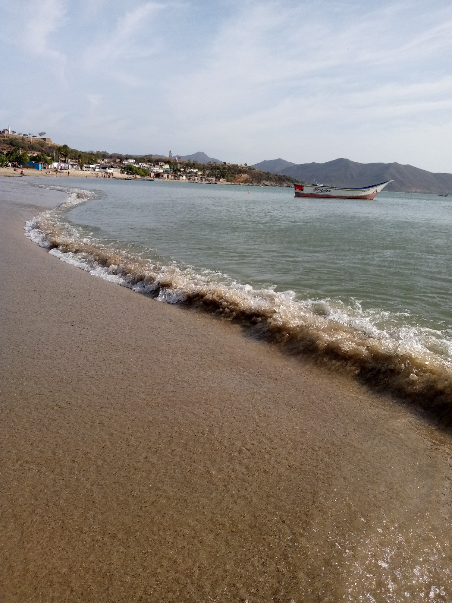 The image captures a coastal scene in Juan Griego, Venezuela. The foreground shows a sandy beach with a wave breaking along the shore. The water appears turbid. In the midground, a white and red boat is anchored offshore with the name "El Ratza" visible on its side. The background features a settlement along the coast, with buildings and structures stretching towards the base of hills and mountains. The sky is partly cloudy.