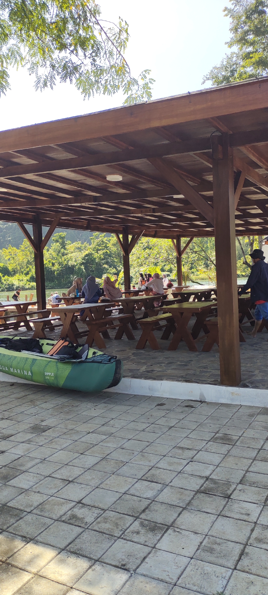 This is an eye-level, outdoor, vertical shot of a covered wooden pavilion with picnic tables in front of a body of water and a wooded hillside. The pavilion has a slatted wooden roof and is supported by wooden posts. Several groups of people are seated at the picnic tables, some appearing to be eating or socializing. The ground within the pavilion and extending outwards is paved with square tiles. To the left of the pavilion, on the tiled ground, rests an inflatable kayak that is green and white. The water appears calm, and the surrounding vegetation is lush and green. The sky is bright and clear, suggesting a sunny day. This scene is located at the Aqua Marina water sports facility in Lamsenia, Aceh, Indonesia.