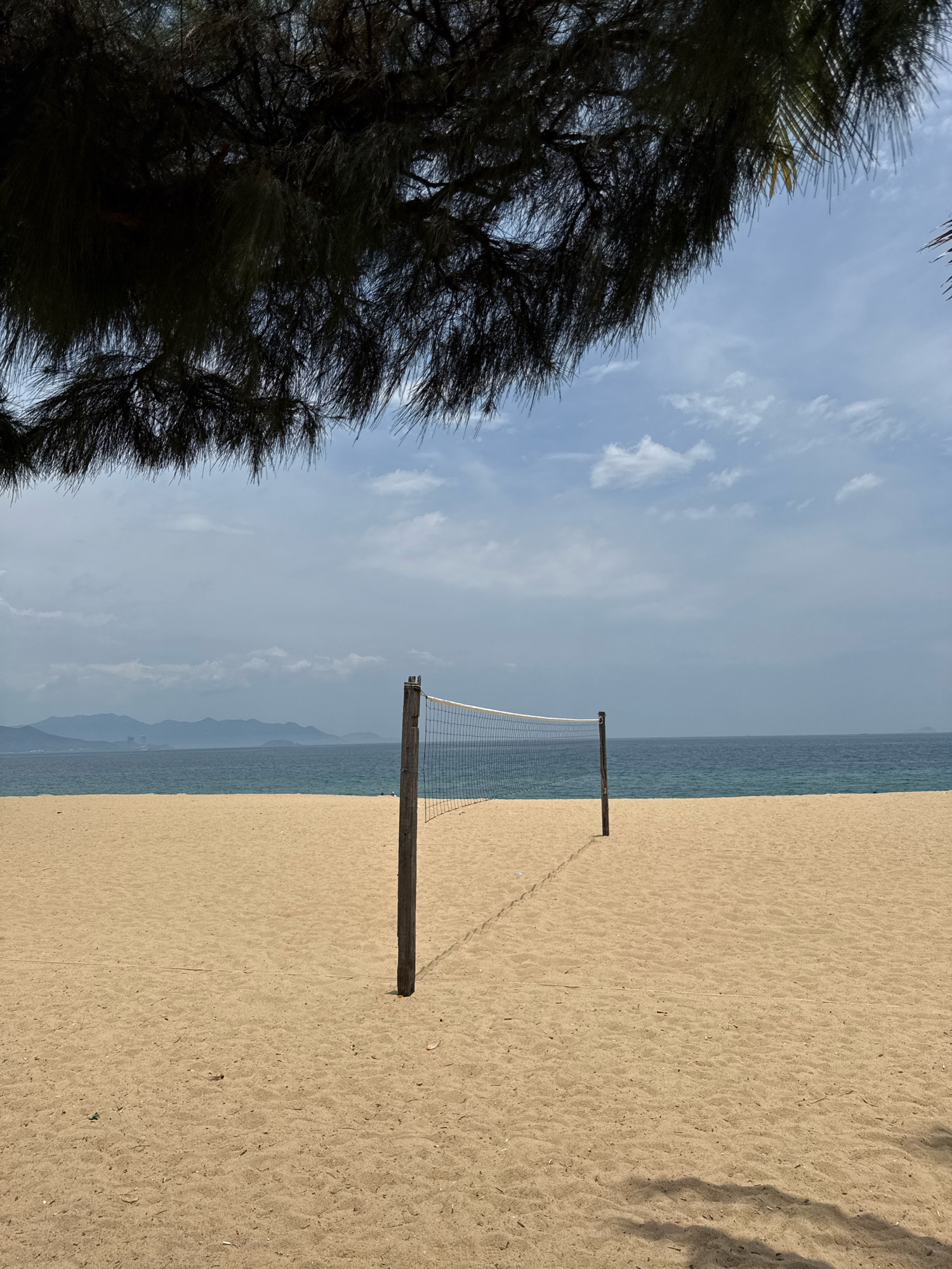 A volleyball net stands on a sandy beach in Nha Trang, Vietnam. The net is positioned between two wooden poles, with the left pole in the foreground and the right pole in the mid-ground. The sand is light brown and appears smooth with some footprints. Beyond the beach, a body of water, likely the ocean, is visible, transitioning from a darker blue closer to the horizon to a lighter turquoise near the shore. In the distance, a mountain range forms the backdrop against a cloudy sky. The image is framed by dark, leafy branches of a tree in the upper portion of the frame, casting shadows on the sand. The overall scene evokes a sense of a tranquil, coastal setting.