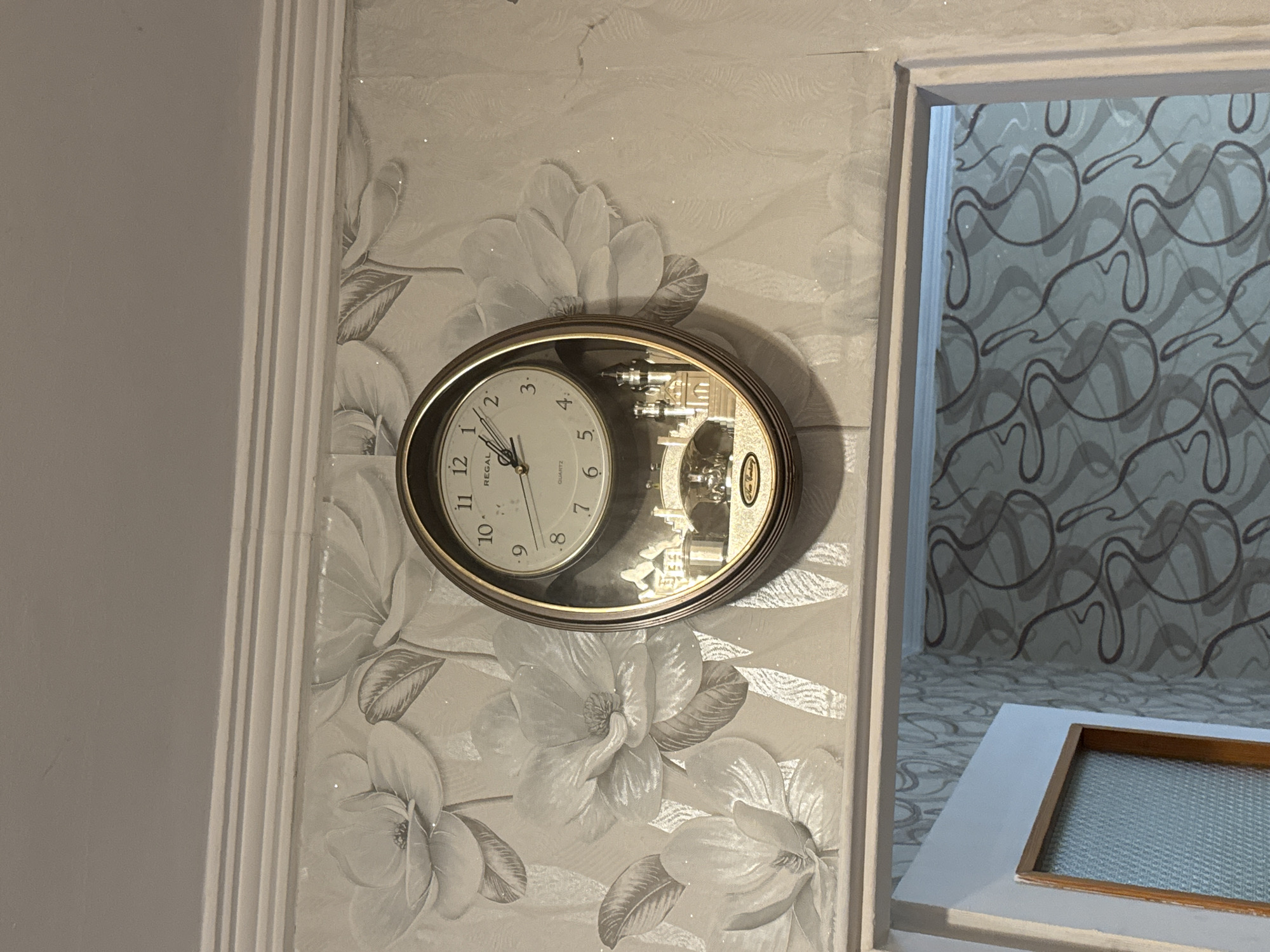 The image depicts an interior scene, likely within a home in Sofia, Bulgaria, focusing on a wall-mounted oval clock. The clock is positioned on a wall covered with light-colored wallpaper featuring large, subtly shimmering grey floral patterns resembling magnolias. To the left of this wallpaper, there's a white, fluted architectural trim piece separating it from a plain grey wall.
The clock itself is an ornamental piece. Its face shows Arabic numerals from 1 to 12, with the time set to approximately 9:48. Visible text on the clock face reads "REGAL QUARTZ." The right half of the clock's interior features an intricate decorative element, appearing as a miniature, stylized cityscape or a complex mechanical scene, reflecting light.
To the right of the clock, an open doorway or archway leads into an adjacent room. The wall visible through this opening is covered with a blue-grey wallpaper adorned with a dark, swirling, abstract pattern. At the bottom of this opening, there is a white frame or ledge with a rectangular wooden inlay surrounding a textured glass panel, possibly a small internal window or a decorative shelf feature. The overall lighting suggests an indoor setting, possibly illuminated by artificial light, given the subtle reflections and glints on surfaces.