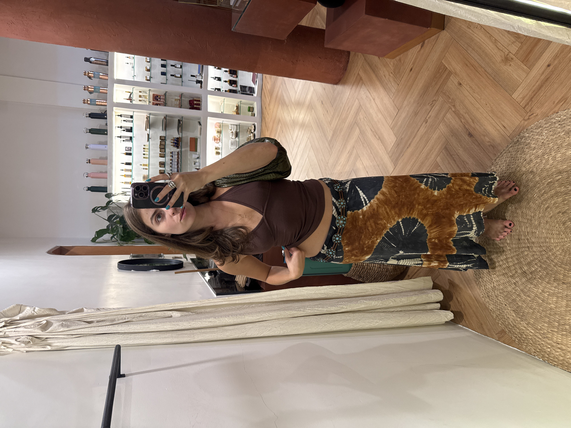 A woman takes a mirror selfie in what appears to be a boutique or retail store in Mengwi, Indonesia. She is wearing a brown halter top, a long patterned skirt with earthy tones, and a textured green purse slung over her shoulder. Her left hand is holding her phone to take the photo, while her right hand is adjusting the hem of her top. She is wearing several rings on her left hand and a necklace. Her toenails are painted black, and she is standing on a woven mat. In the background, shelves are stocked with various items, possibly cosmetics or artisanal products. The flooring is made of light-colored wood laid in a herringbone pattern. A large, rust-colored pillar is visible to the right of the woman.