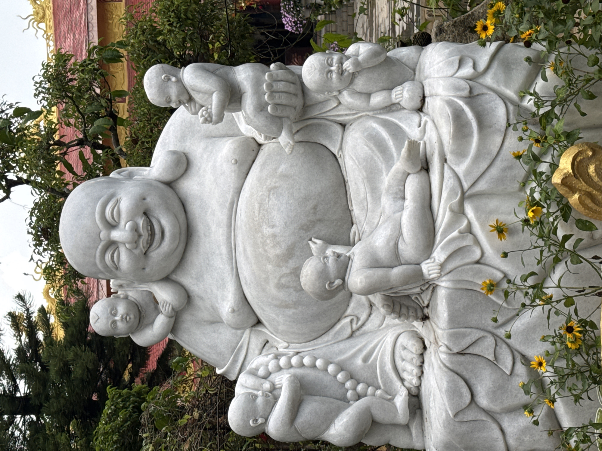 A large, white marble statue of a laughing Buddha is the central focus of the image, situated outdoors in a garden setting. The Buddha is depicted with a prominent belly and a jovial expression, surrounded by several smaller marble figures of babies. One baby sits on the Buddha's shoulder, another on his hand, and others are positioned around his lap and feet, some interacting with a beaded necklace draped around the Buddha. The statue is placed on a stone platform, and the foreground is filled with green foliage and yellow wildflowers. In the background, a red-roofed building with gold architectural details is visible, partially obscured by lush green trees. The overall scene suggests a temple or a garden dedicated to Buddhist imagery in Lâm Viên - Đà Lạt, Vietnam.