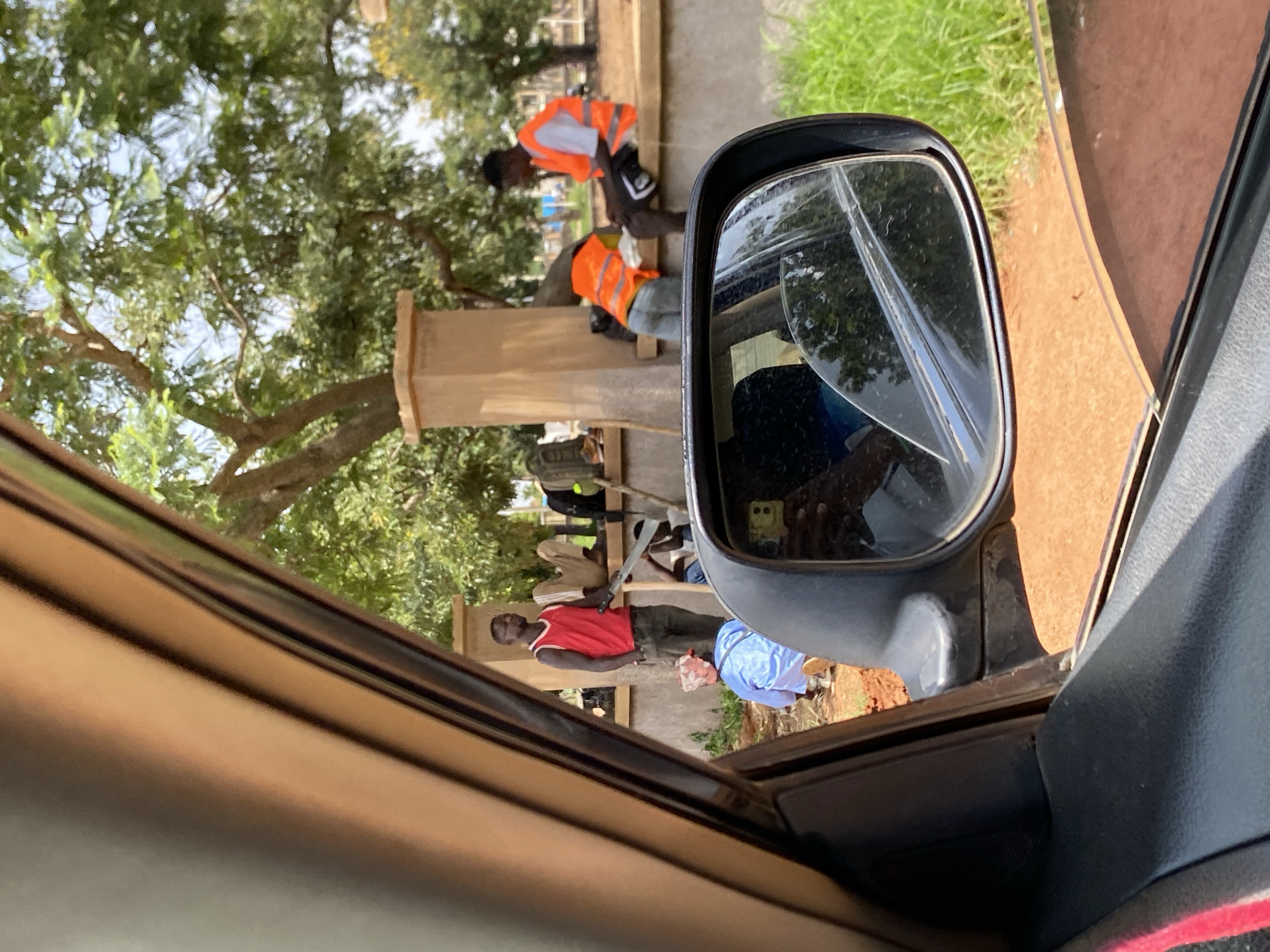 In Bar Jirgi Summa, Nigeria, a person wearing a red sleeveless shirt and dark pants stands holding a machete. Another person, dressed in a blue garment, is partially visible in the foreground. Two individuals are seated on a low wall, one wearing an orange safety vest and the other also in an orange safety vest. The environment suggests an outdoor setting with trees and grassy areas, observed from within a vehicle looking out through the side window and rearview mirror. The ground appears to be red soil.