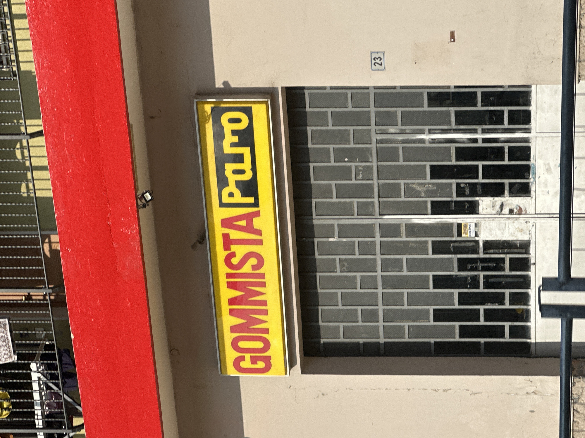 A vertical, rectangular yellow sign with red lettering hangs on a beige building wall. The sign reads "GOMMISTA Pa.ro". Next to the sign is a window with a grey metal security grate. Above the window, a small blue and white number "23" is displayed on the wall. To the left of the sign and window, a bright red, flat surface is visible, likely a painted wall or door. In the background, to the left of the red surface, metal railings and a chain-link fence suggest an outdoor area. The lighting and shadows indicate it is daytime, likely sunny. The scene appears to be a storefront or business in Rudiano, Italy, given the context. There are no people visible in the image.