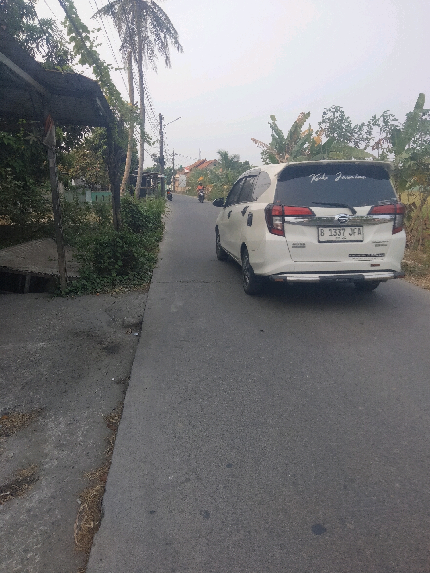 A white minivan, license plate B 1337 JFA, is parked on the right side of a paved road in Suka Sari, Indonesia. The minivan has a black sticker on its rear window that reads "Keiko Jasmine" in white script. The road is lined with vegetation, including palm trees and banana trees, and on the left side of the road, there is a structure with a corrugated metal roof. Two motorcycles are visible further down the road, one behind the other, with riders on them. The sky is overcast.