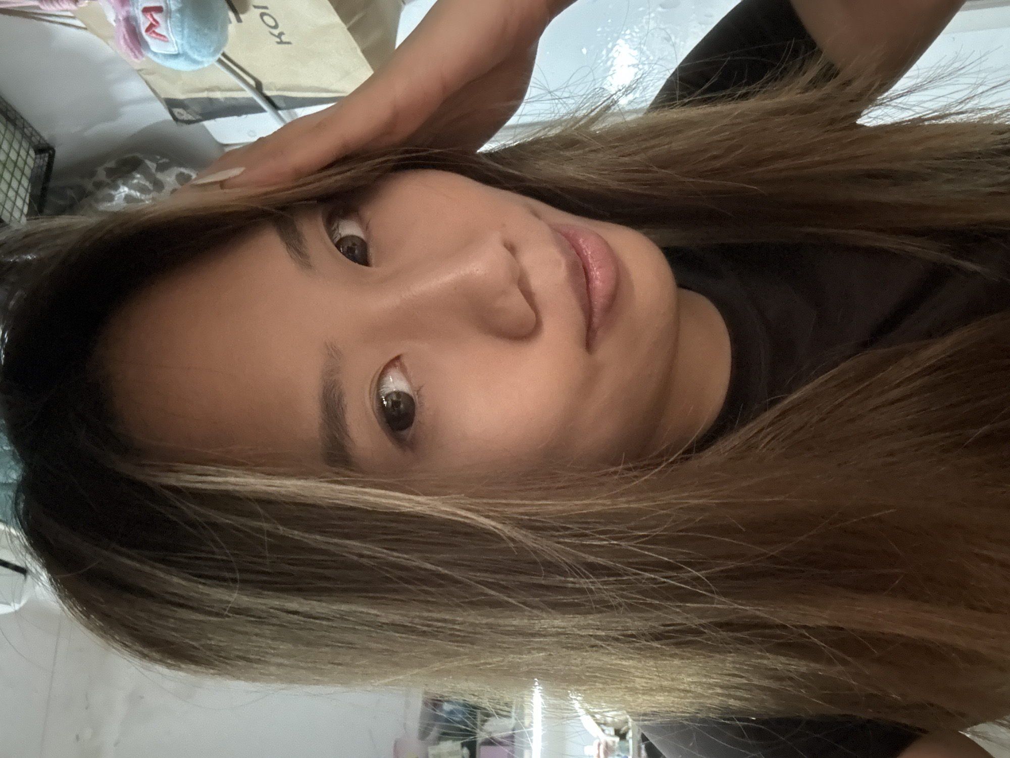 A close-up, angled, indoor portrait shows a young Asian woman with shoulder-length, light brown hair with blonde highlights. Her hand is on her forehead, as if she is pushing back her hair. She is wearing a dark-colored top. Her expression is neutral, with brown eyes looking directly at the camera. A shopping bag and a small, stuffed animal are visible in the background, along with glimpses of a cluttered, indoor environment. The location is presumed to be indoors within the Nan'an District, China.