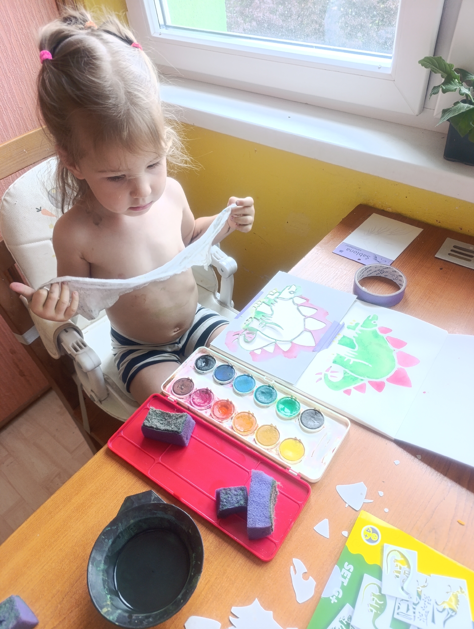 A young Caucasian child with blonde hair tied into pigtails sits in a high chair at a wooden table, engaged in an art project. The child, wearing striped shorts, holds a wet wipe in their hands, appearing to clean their torso. The table is covered with art supplies, including a watercolor paint set with various colors, sponges, and stencils. Two drawings of dinosaurs are visible, one partially completed with green paint. A roll of tape and a small plant in a pot are also on the table. The background features a window with a view of greenery and a yellow-painted wall. The scene suggests a creative activity taking place in a home environment, potentially in Olomouc, Czechia.
