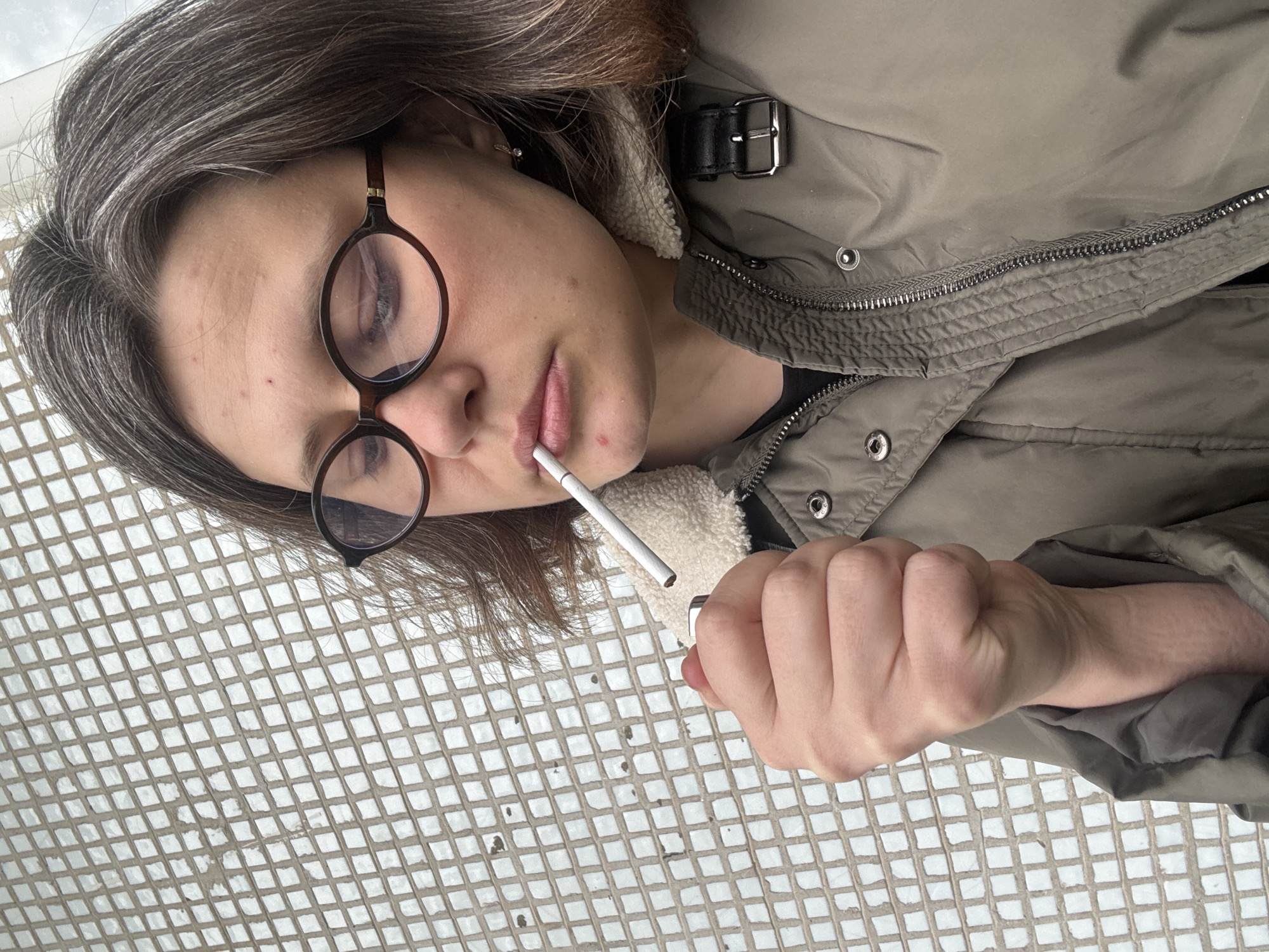 A woman with graying hair and round glasses holds a lit lighter to a cigarette in her mouth. She wears a light green jacket with a white furry lining and is standing on a white tiled surface. The exact location within Chaikovsky, Russia, cannot be determined from the image alone.