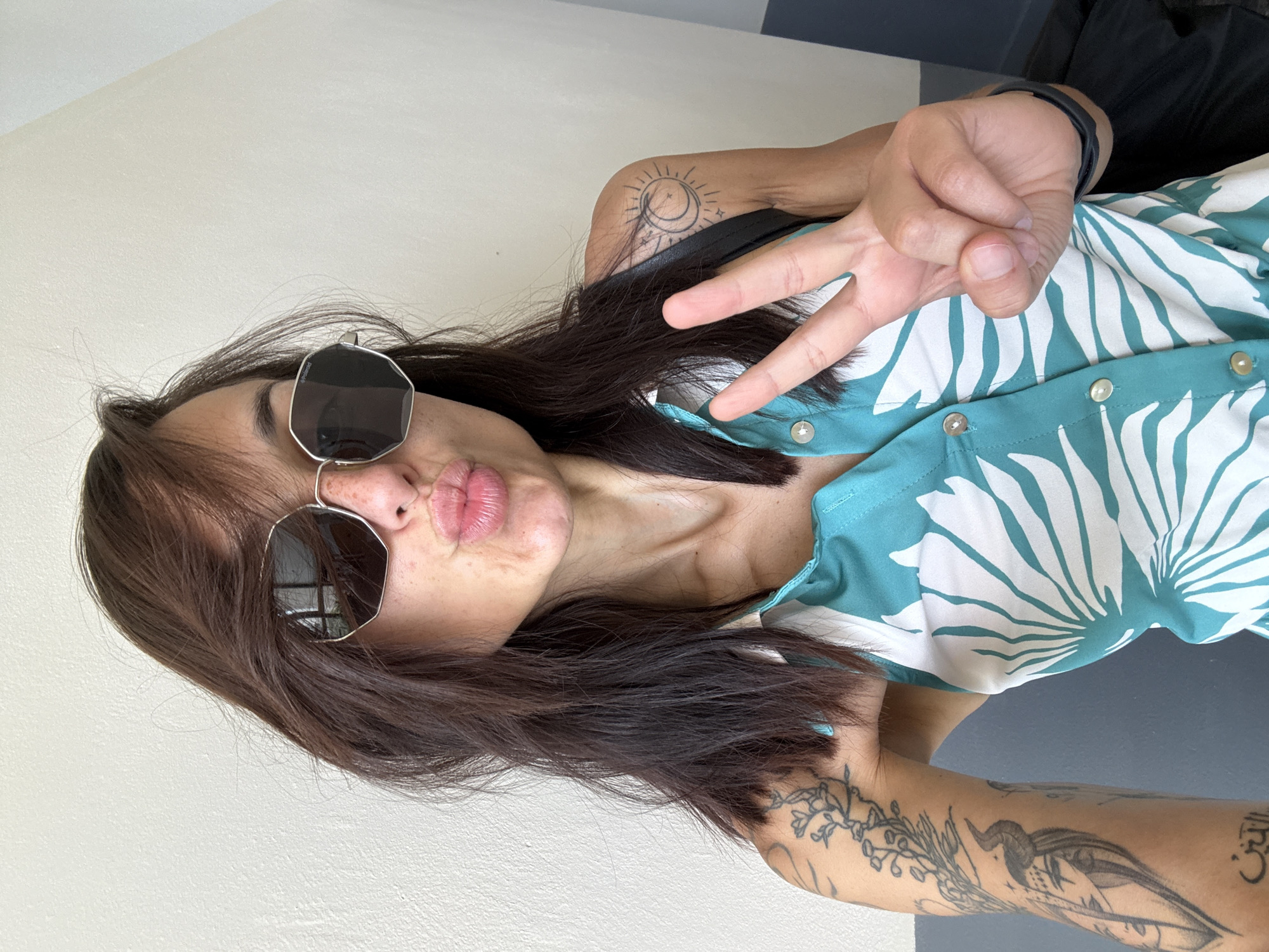 The media file shows a person taking a selfie. The person is wearing sunglasses, a teal and white patterned shirt, and has multiple tattoos on their arms and shoulder. They are making a peace sign with their right hand and puckering their lips. The background appears to be indoors, with a light-colored wall on the left and a darker surface on the right. The lighting suggests it is daytime.
