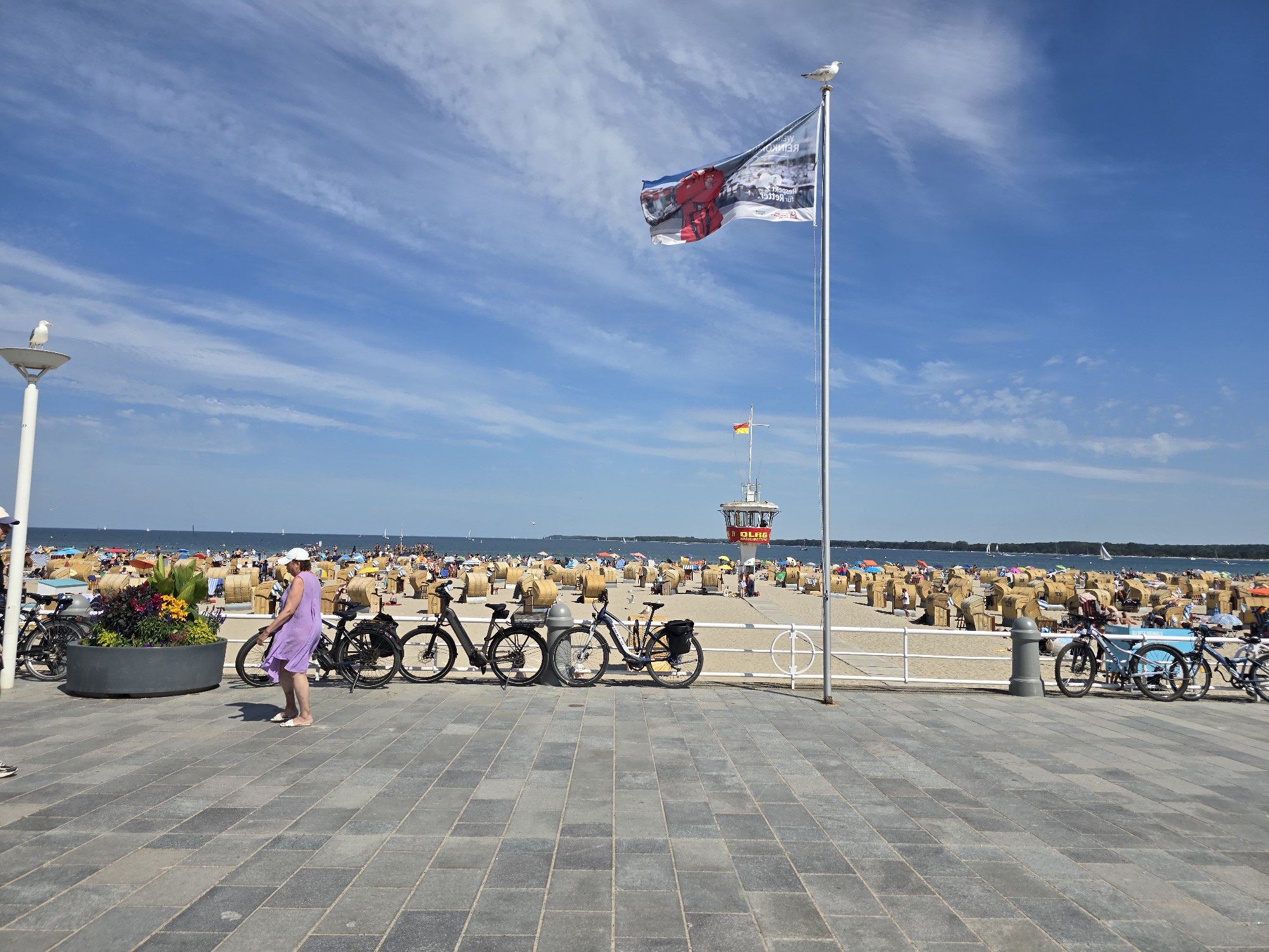 This image depicts a sunny day on a beach promenade in Travemünde, a district of Lübeck, Germany. The foreground shows a paved walkway with several bicycles parked along a white fence. A woman in a purple dress and white hat is walking on the promenade, and a decorative planter with colorful flowers sits near the bicycles.
Beyond the fence, the beach is filled with rows of wicker beach chairs under umbrellas, indicating a popular recreational area. In the distance, the Baltic Sea stretches out under a bright blue sky with scattered clouds. A lifeguard tower, marked with "DLRG," stands prominently on the beach, a common sight at German beaches. A flag flutters from a tall pole on the promenade, and seagulls are visible perched on a lamppost and the flagpole. The scene conveys a lively summer atmosphere.