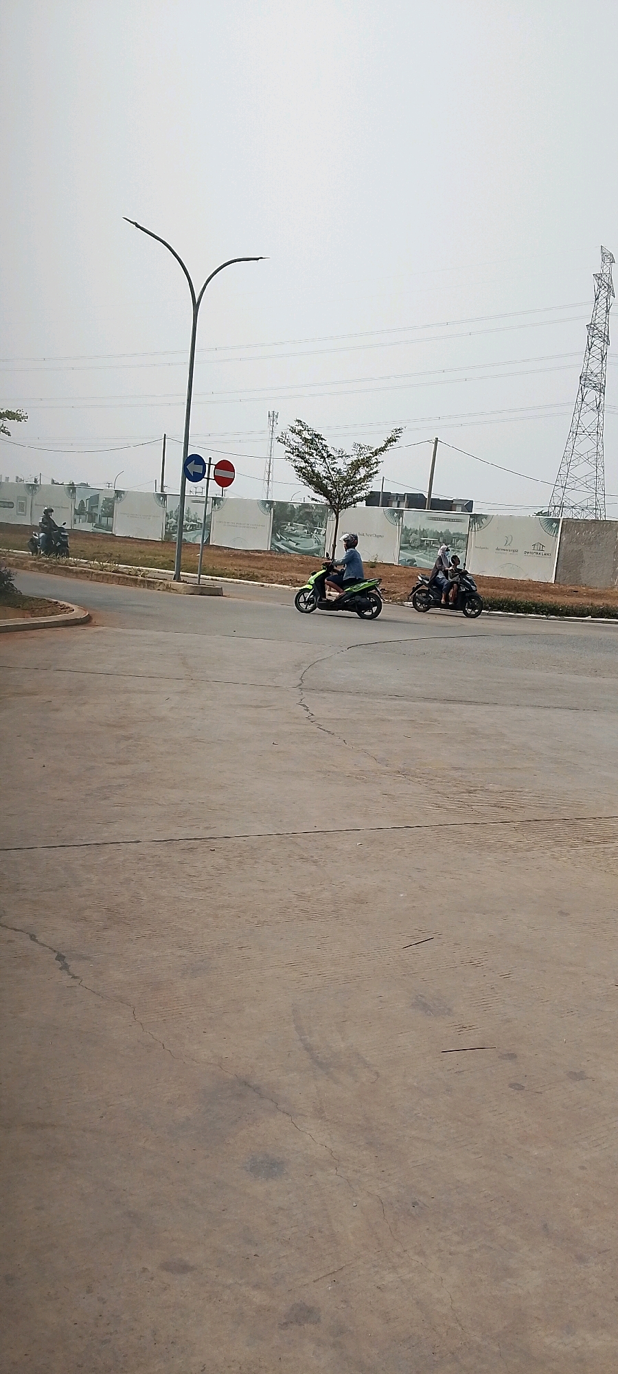 This image depicts a street scene in the city of Satriamekar, Indonesia. The perspective is from the ground level, looking slightly upwards towards the sky, which is overcast and pale. In the foreground, a concrete road surface is visible, showing signs of wear and tear with cracks and some darker patches.
Further down the road, several motorcycles are in motion. On the right side, a green motorcycle is ridden by a person wearing a helmet and a blue shirt. Behind them, another black motorcycle carries two individuals, both wearing headscarves and helmets. To the left, a third person on a motorcycle is visible in the distance, also wearing a helmet.
Along the side of the road, a sidewalk separates the road from a construction site. A tall street light with a distinctive curved design stands on the left side of the road. A traffic sign, featuring a blue arrow pointing left and a red circle with a white horizontal bar, is also present. Behind the sidewalk, a large fence or hoarding displays promotional images of housing developments. In the background, a tall electrical pylon and power lines are visible against the sky, along with other distant structures and what appears to be trees. The overall environment suggests an urban or suburban developing area.