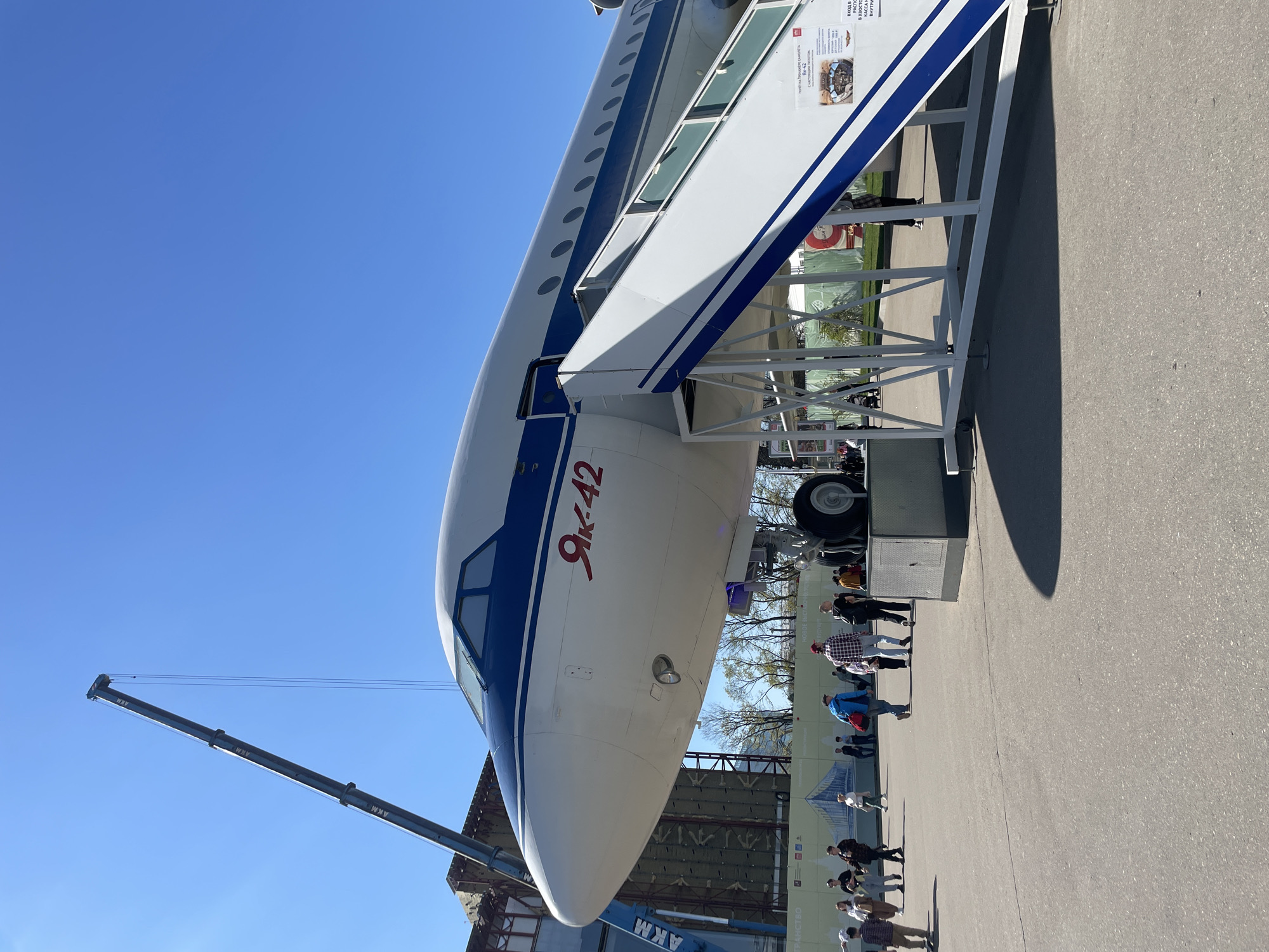 The image depicts the nose section of a Yakovlev Yak-42 aircraft, a medium-range passenger jet, prominently marked with "Як-42" in Cyrillic script. A white boarding staircase with blue accents is attached to the aircraft, indicating possible public access. In the background, several people are visible on the ground, likely visiting the exhibit. A crane arm extends into the frame on the left side of the aircraft. The scene is situated outdoors on a paved surface under a clear blue sky. Based on the Cyrillic signage and aircraft type, the location is likely within the city of Moscow, Russia, potentially at an aviation museum or exhibition.