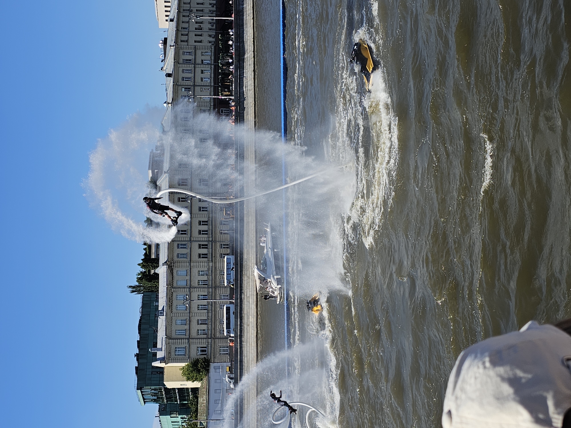 This image depicts a sunny day on a wide river in Moscow, Russia, likely the Moskva River. In the foreground, the back of a person's head wearing a light-colored baseball cap is visible, looking out at the water. The main action unfolds on the river with individuals engaged in flyboarding.
One flyboarder, dressed in dark attire with a helmet, is suspended high in the air, propelled by streams of water ejected from a device attached to their feet. They are connected by a hose to a watercraft or pump system, creating a dramatic spray of water.
Further out on the water, two yellow jet skis are active. One is creating a significant wake, and another is partially submerged, with a person visible riding it. A white motorboat with several passengers is also present on the river, traveling in the background.
The river is bordered by a stone embankment, and beyond that, a series of buildings line the cityscape. These structures appear to be multi-story, with architectural details suggesting they are older buildings, some with multiple windows and a lighter-colored facade. Modern buildings with darker facades and visible glass are also present in the background, indicating a mix of architectural styles. The sky is clear and blue. The overall scene is one of outdoor recreation and urban scenery.