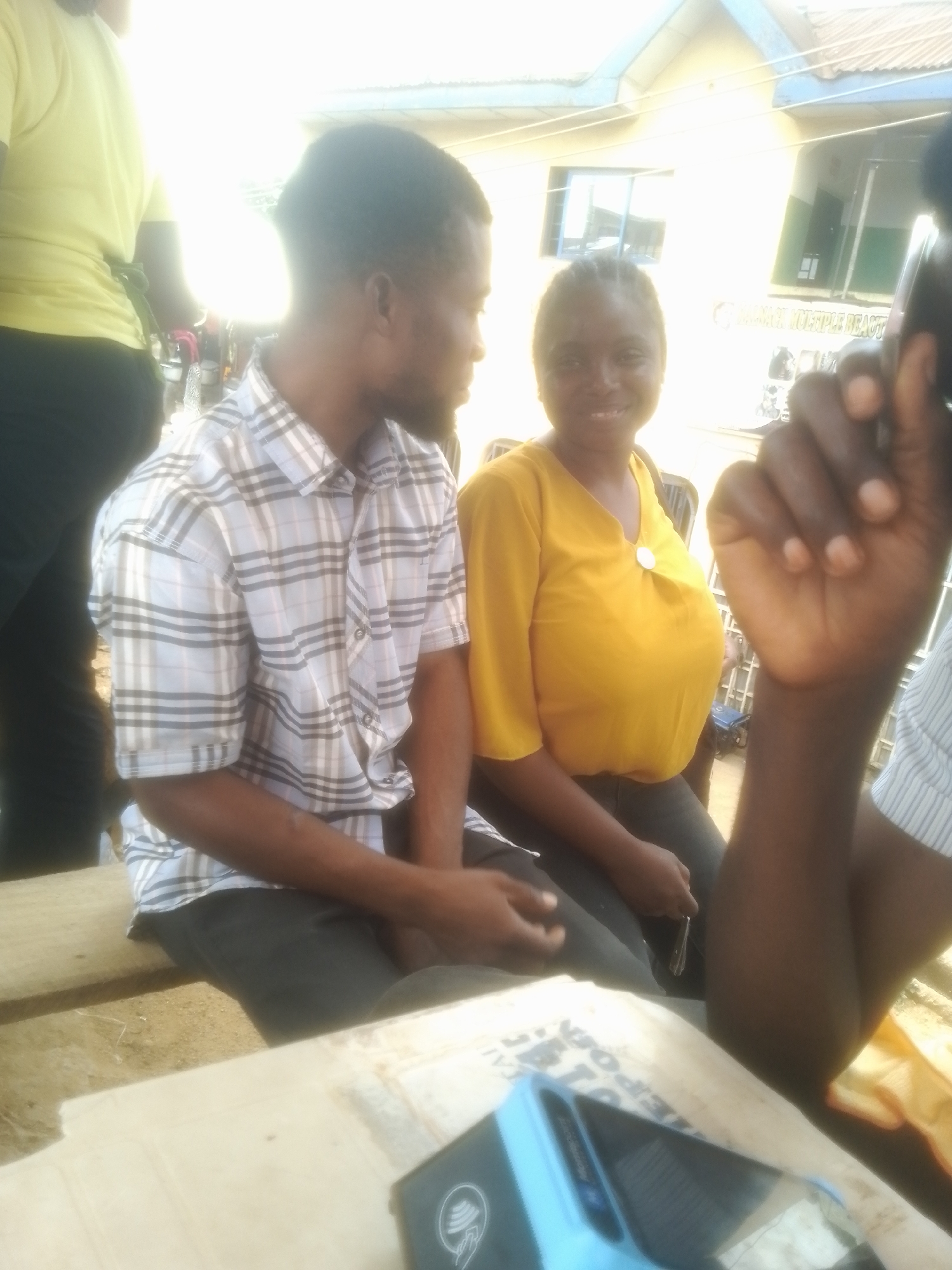 Two individuals are seated on a bench outdoors, with a building in the background. A person with dark skin and short hair, wearing a plaid shirt, is on the left. To their right, a person with dark skin and braided hair, wearing a yellow v-neck shirt, is smiling. In the foreground, a hand is visible, and a blue electronic device with a screen is on a surface. The environment appears to be a public area, possibly a market or waiting area, given the presence of the bench and the building. The lighting suggests it is daytime.