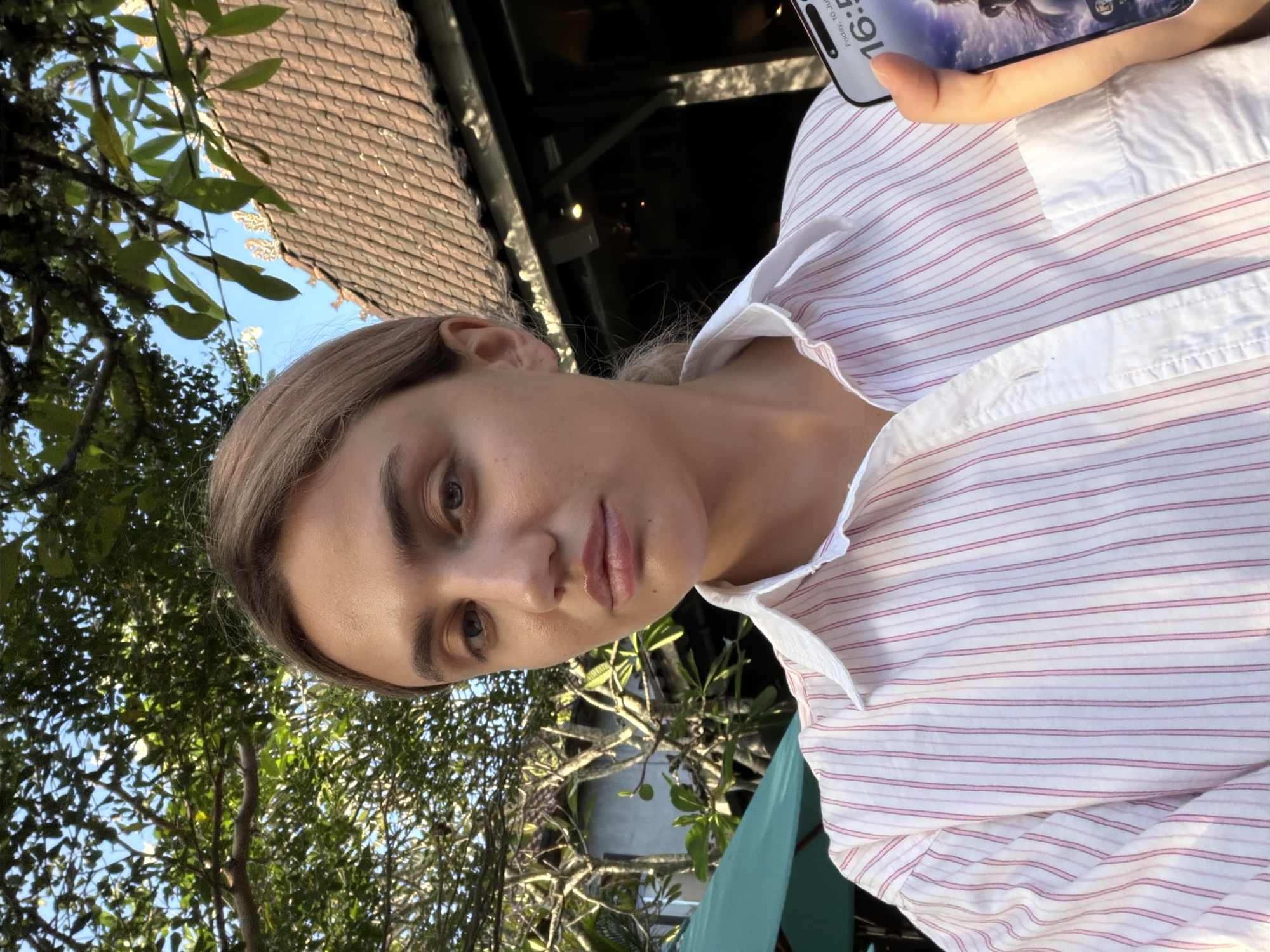 A young woman with blonde hair pulled back is taking a selfie outdoors in what appears to be a tropical location. She is wearing a white collared shirt with thin pink vertical stripes. The background shows lush green trees, a glimpse of a tiled roof building, and a blue sky. In her right hand, she is holding a smartphone, which displays the time as "16:53" and the date as "Friday, 10 July". The lighting suggests it is late afternoon. The overall impression is of a relaxed, candid moment captured in a warm climate, possibly a resort or cafe in Ubud District, Indonesia, given the architectural style and foliage.