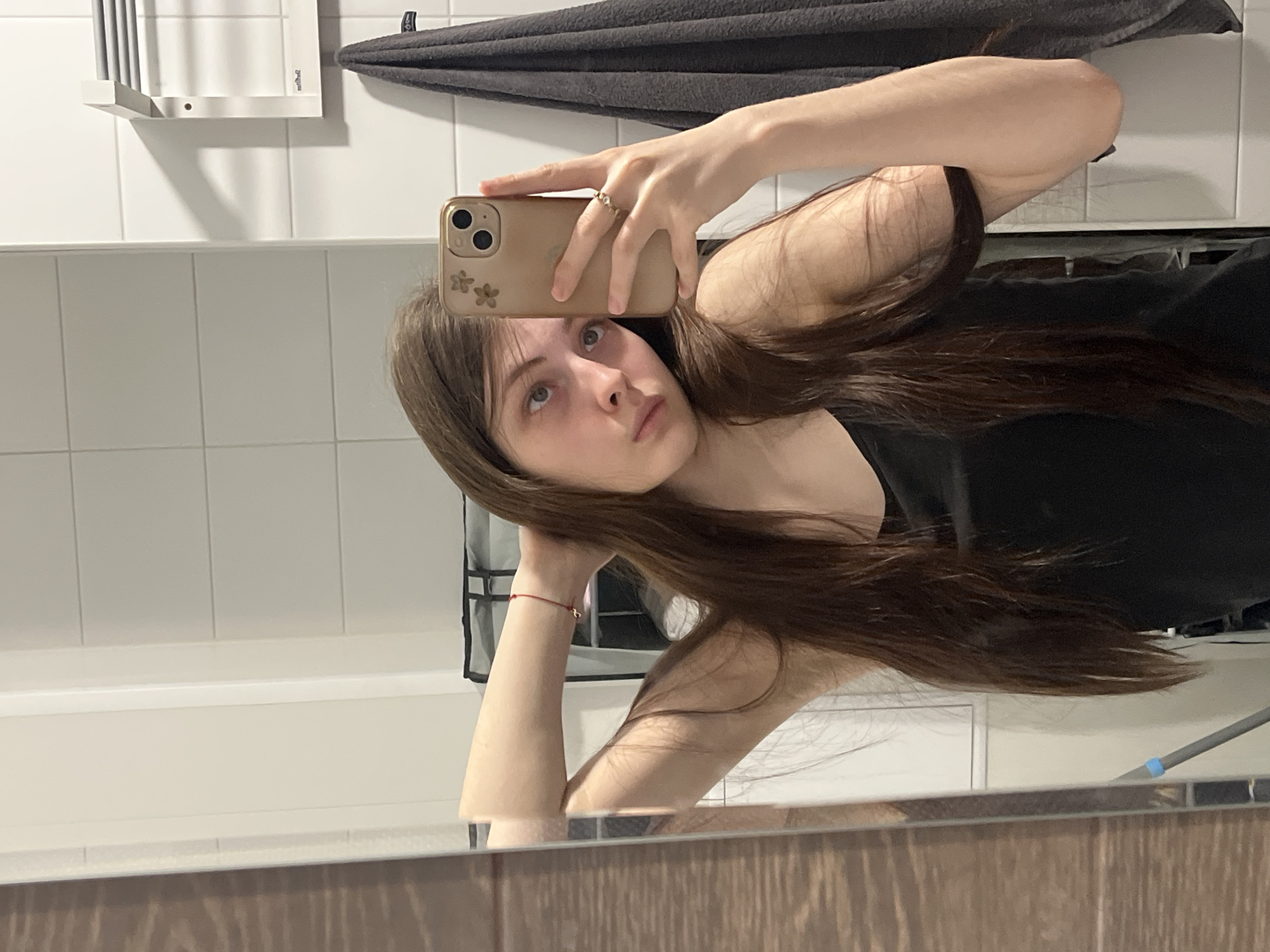 A young woman with long brown hair is taking a selfie in a bathroom mirror. She is wearing a black top and a ring on her left hand. The bathroom has white tile walls with a grey towel rack and a small white shelf visible in the background. The vanity area of the bathroom has brown paneling. The image does not contain any information to determine if it's in Saint Petersburg, Russia.